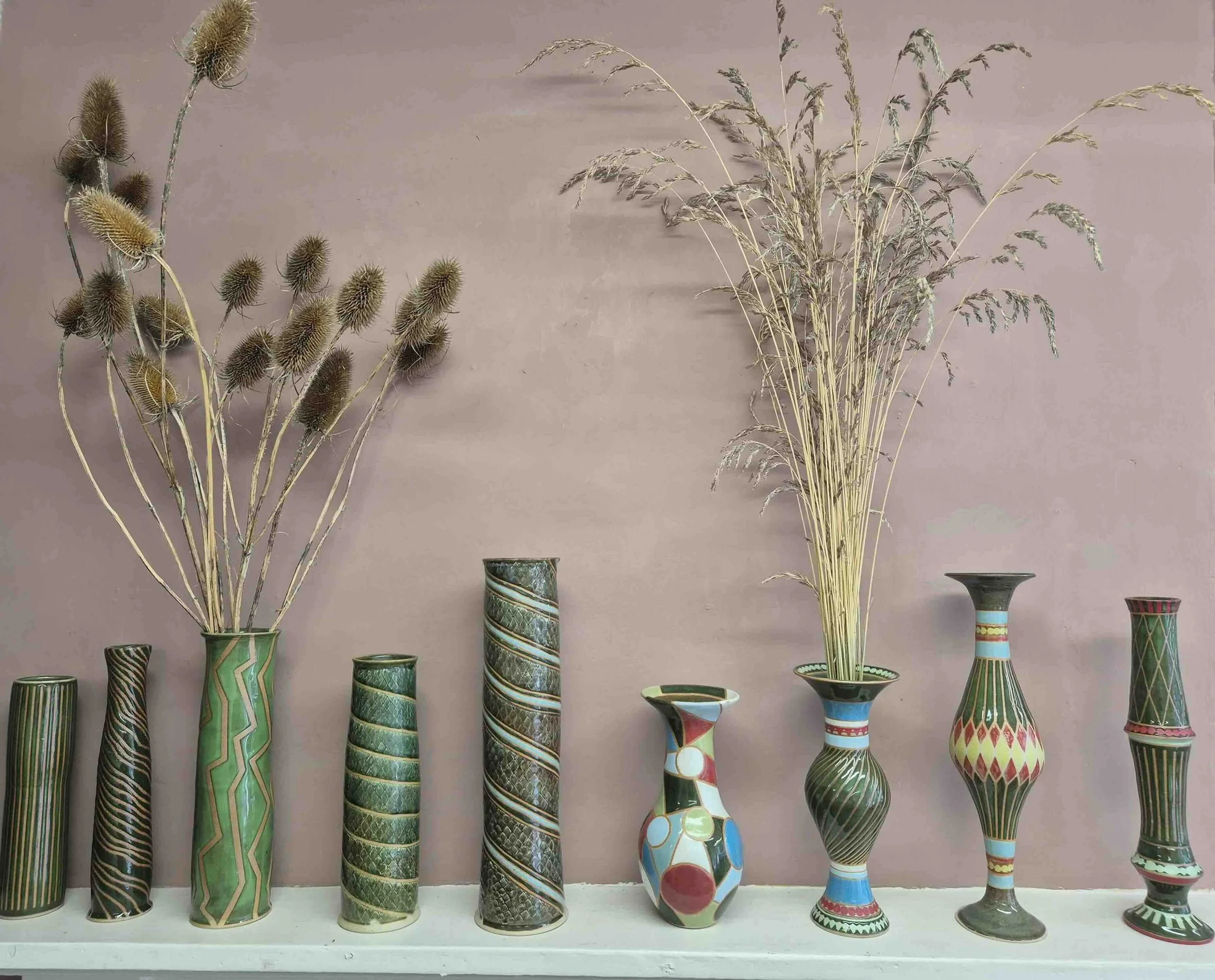 Decorative vases by Emily Clarke with dried plants and flowers arranged in a lineup on a shelf against a pink wall.