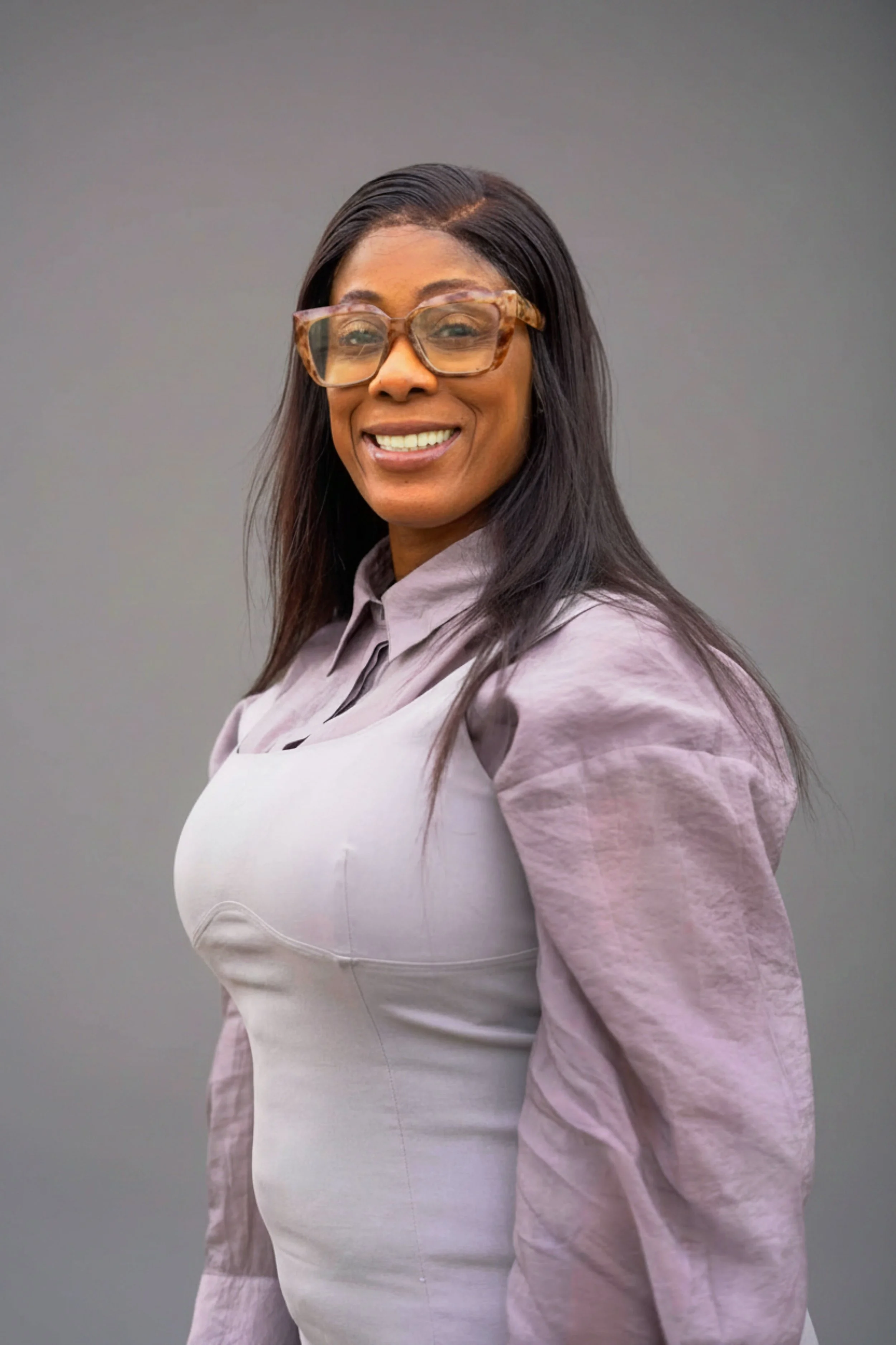 Jennie Bourne smiling with short black hair, glasses, and a tattoo on her chest, dressed in black, in an indoor setting.