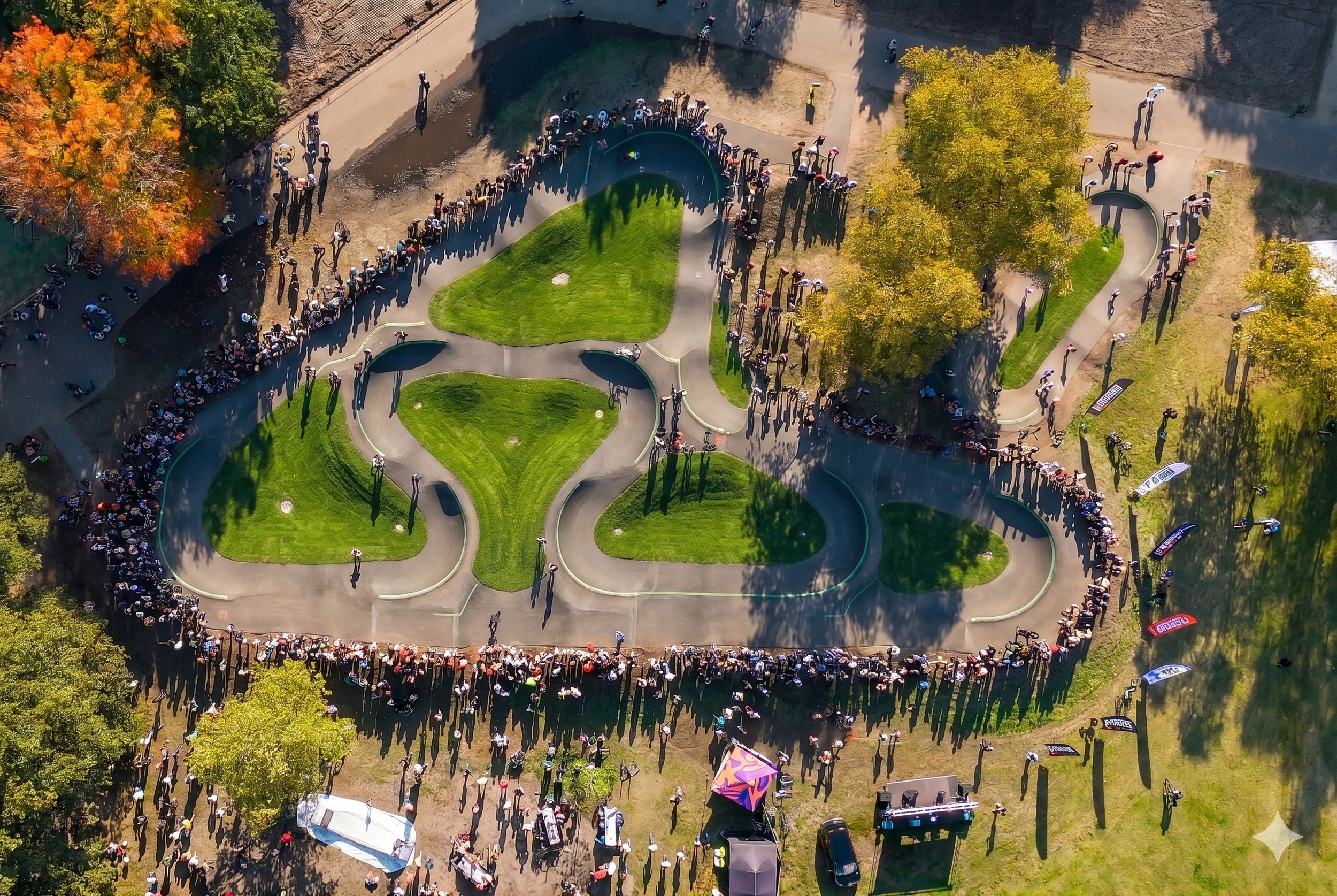 Luchtfoto van een park met een fietspad en een pumptrack waar veel mensen aanwezig zijn, omringd door herfstbomen met oranje, geel en groen blad.