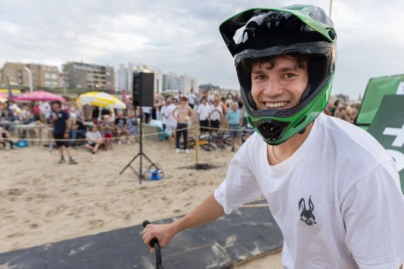 Man met fietsmasker op op het strand, omringd door een menigte onder een bewolkte hemel.