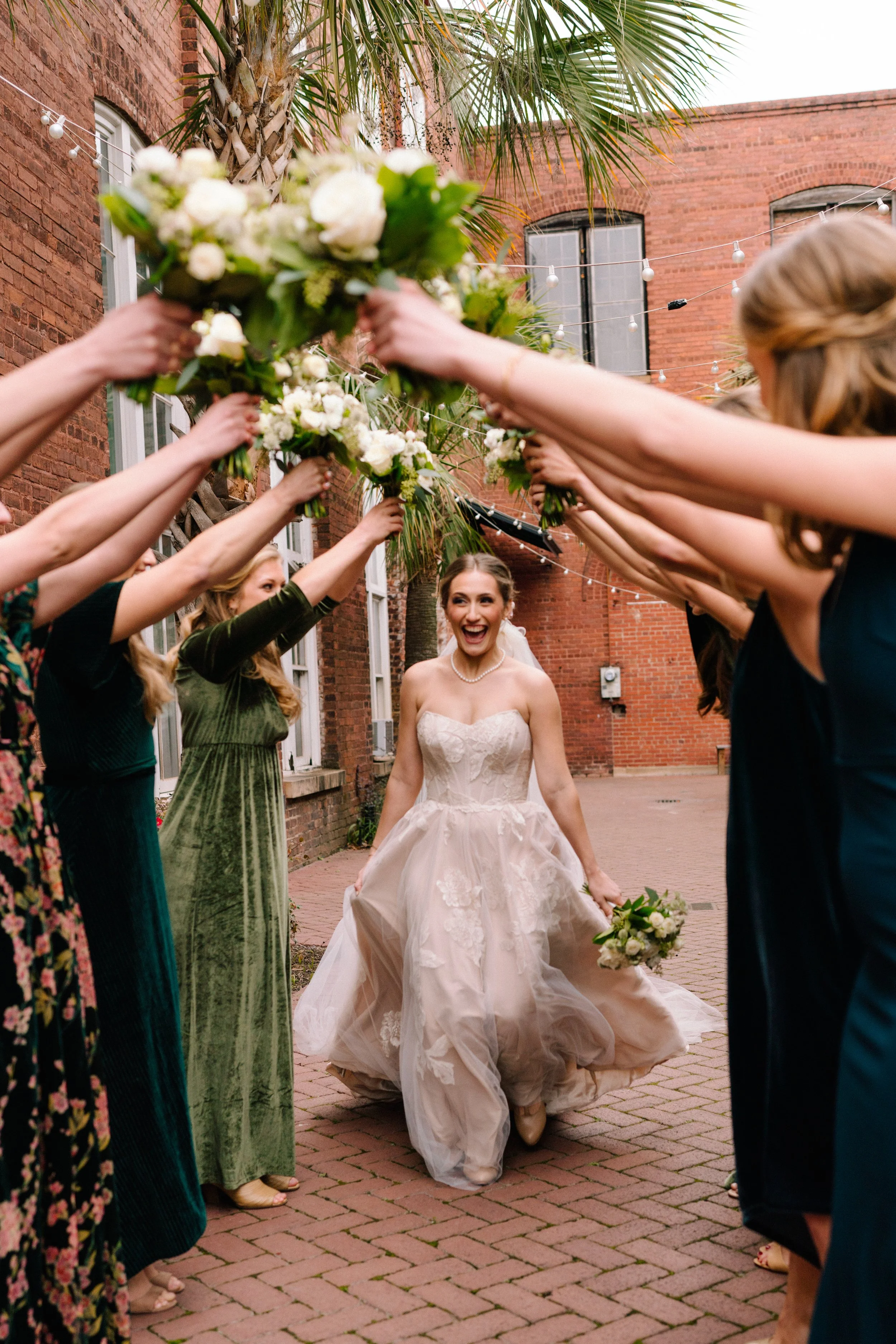 A medium shot of Emily walking in between her bridesmaids in a "tunnel" formation. They hold their bouquets to form an archway.