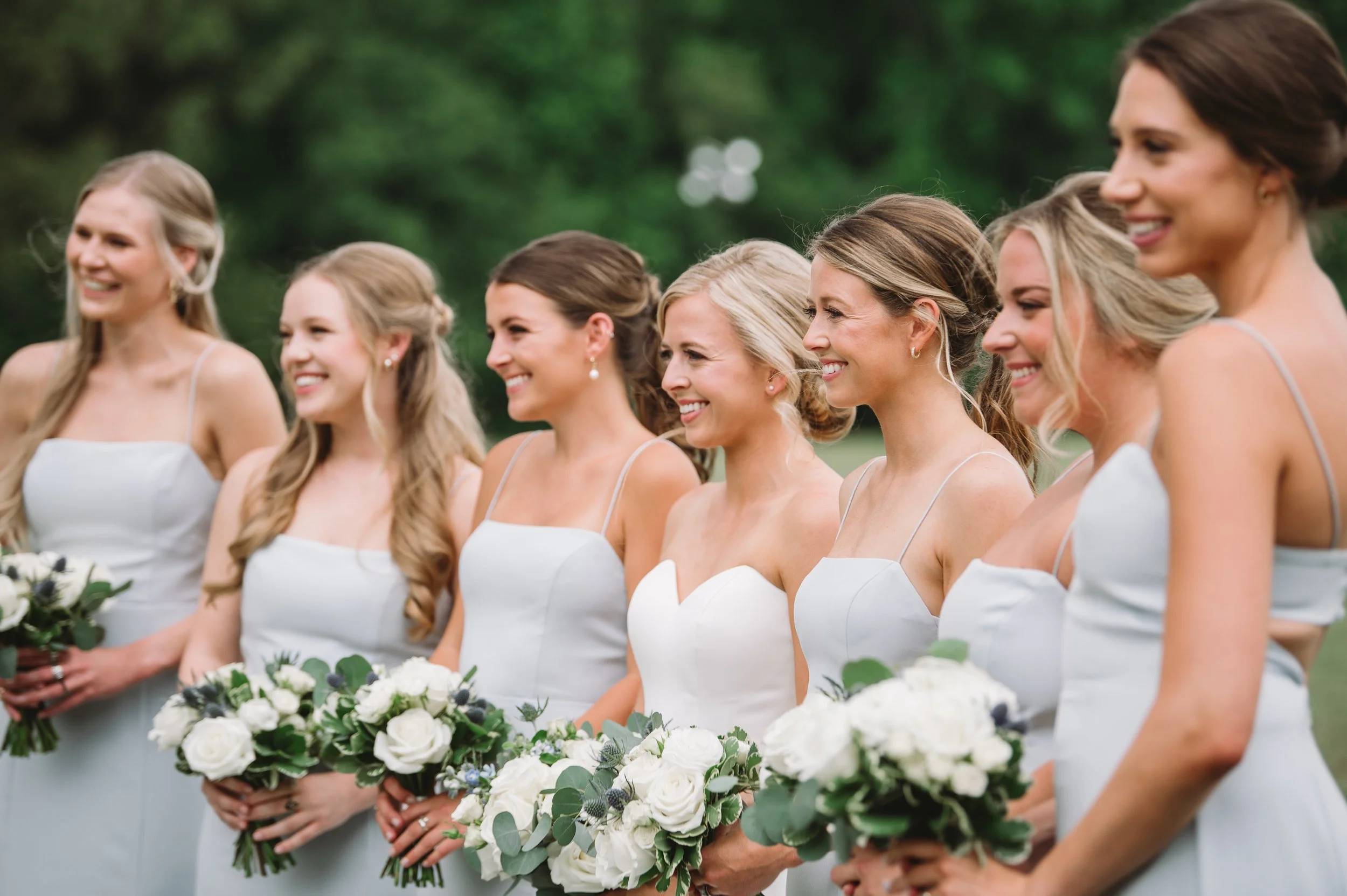 A picture of Callie and her bridesmaids smiling for a different photographer. They are facing left and are holding their bouquets. The bridesmaids are wearing light blue grey dresses.