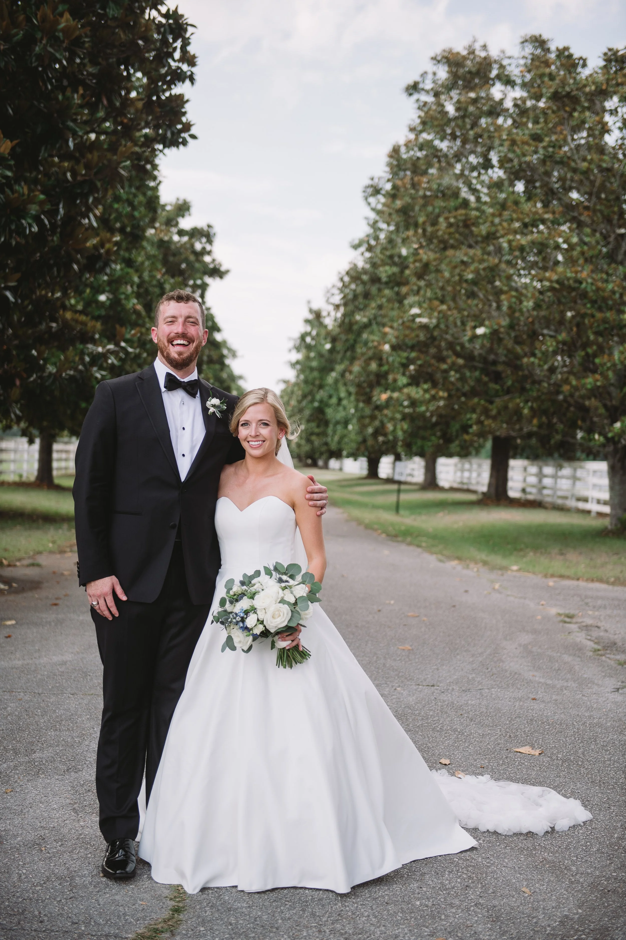 A portrait of Callie and John taken on a road. There are trees and a white fence in the background.