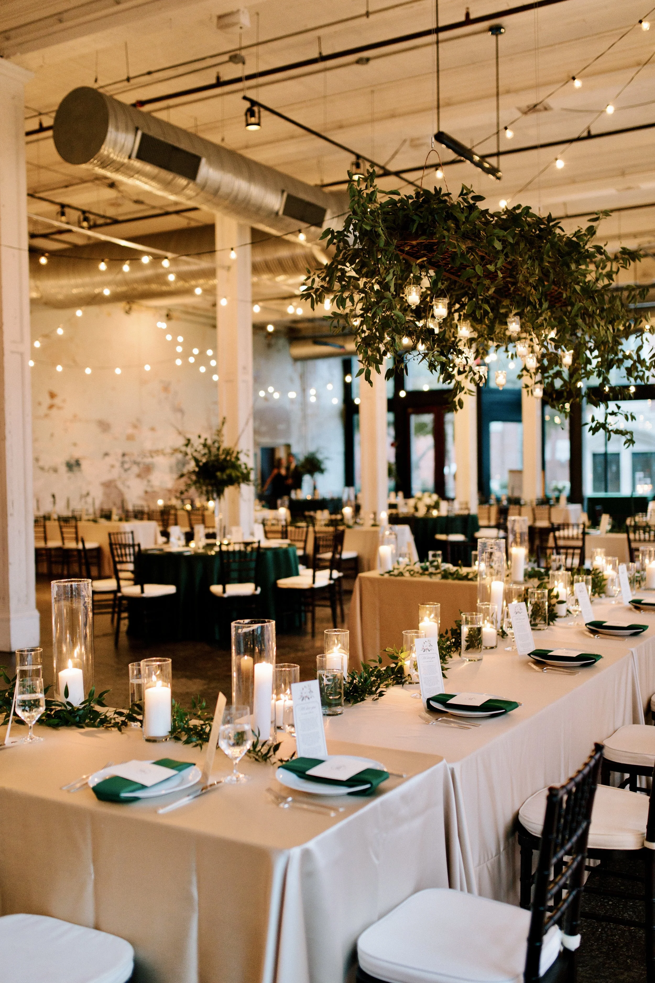 A shot of the reception hall showing a large hanging green vine statement piece and the party's table. There are place settings and white candles and vines as decor.