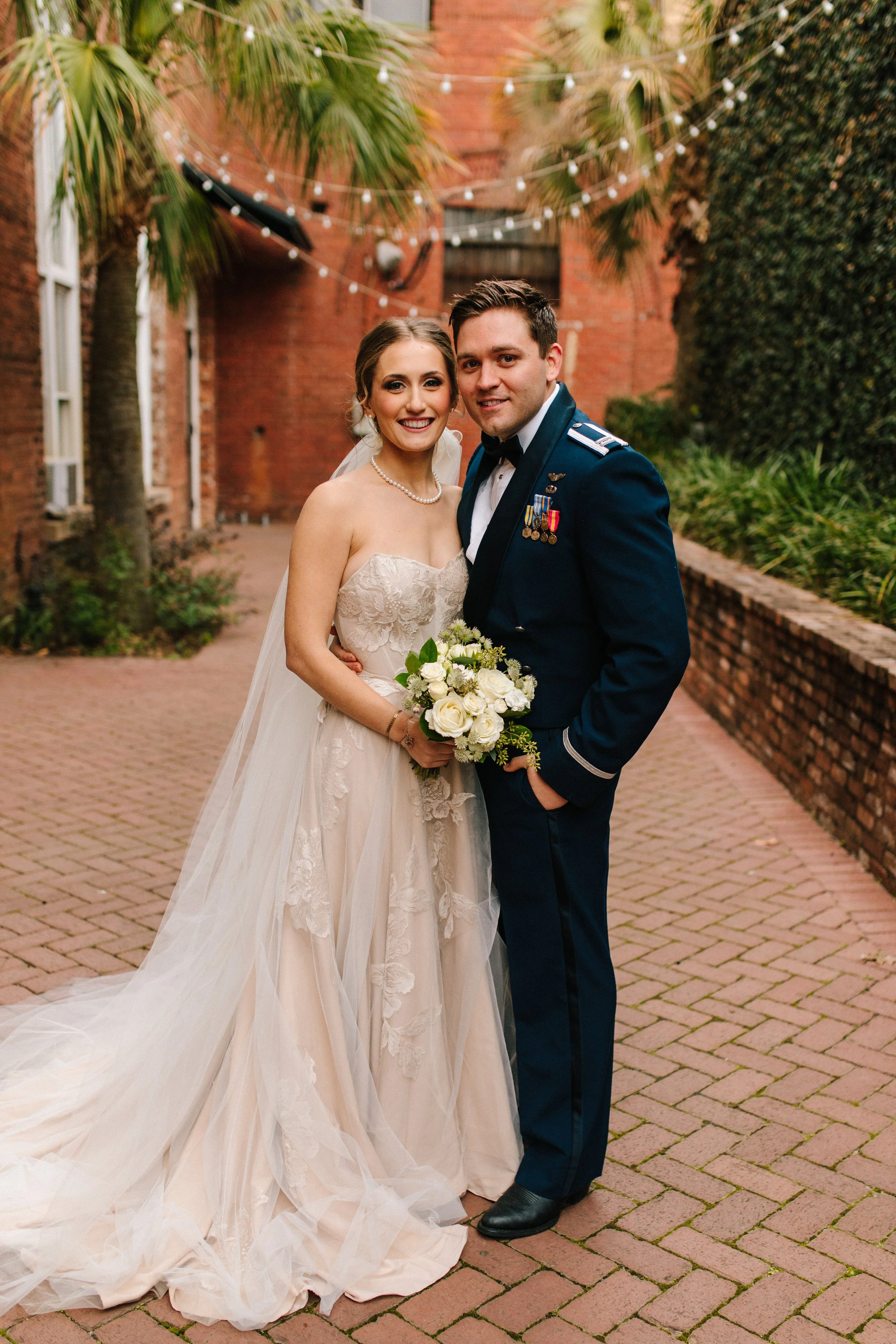 A traditional portrait of Emily and Hunter on a brick patio. She holds her bouquet and there are string lights and plants in the background