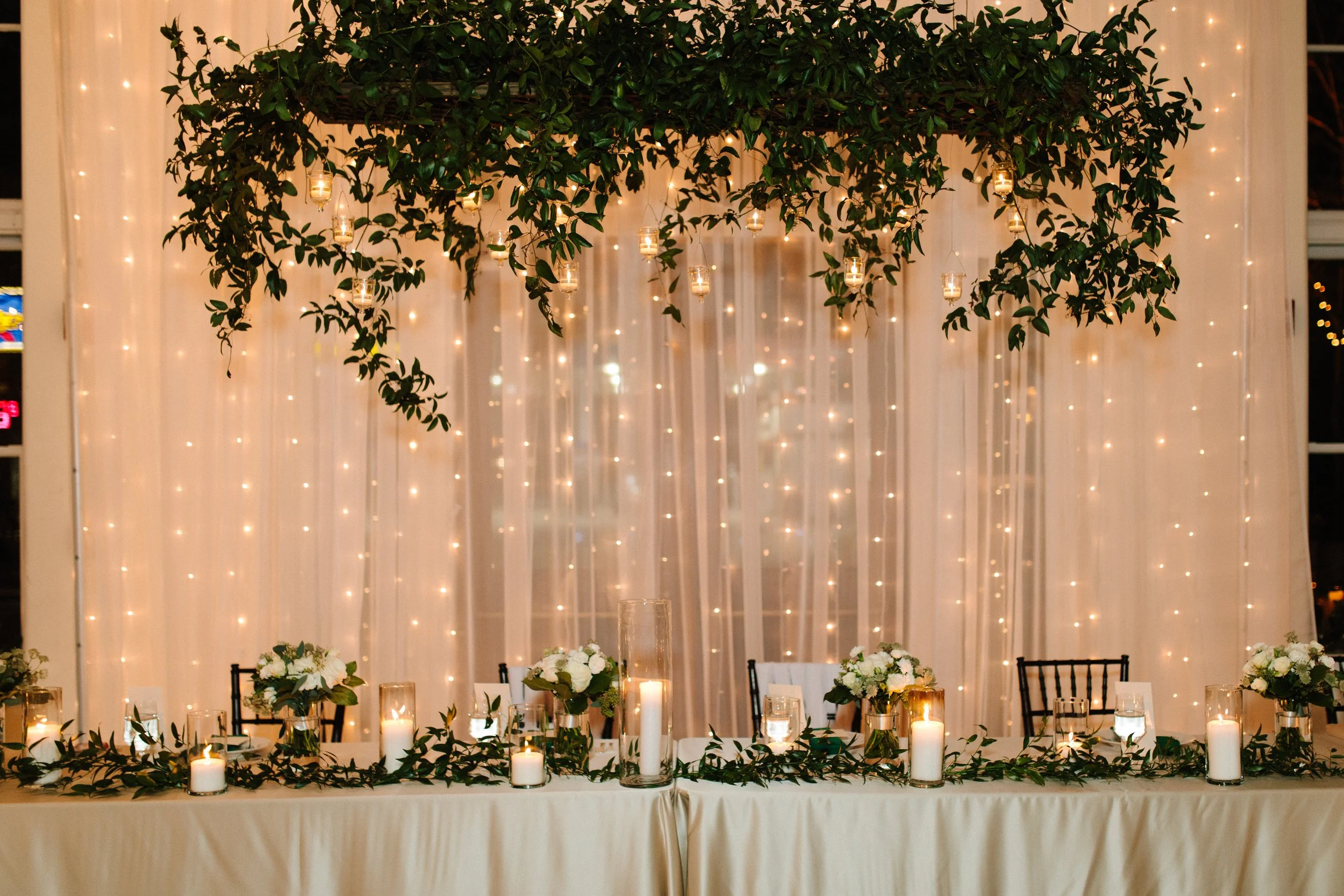 A straight on shot of the party's table. There is a large hanging floral/vine arrangement above and drapes and string lights in the background. On the table there are vines and candles.