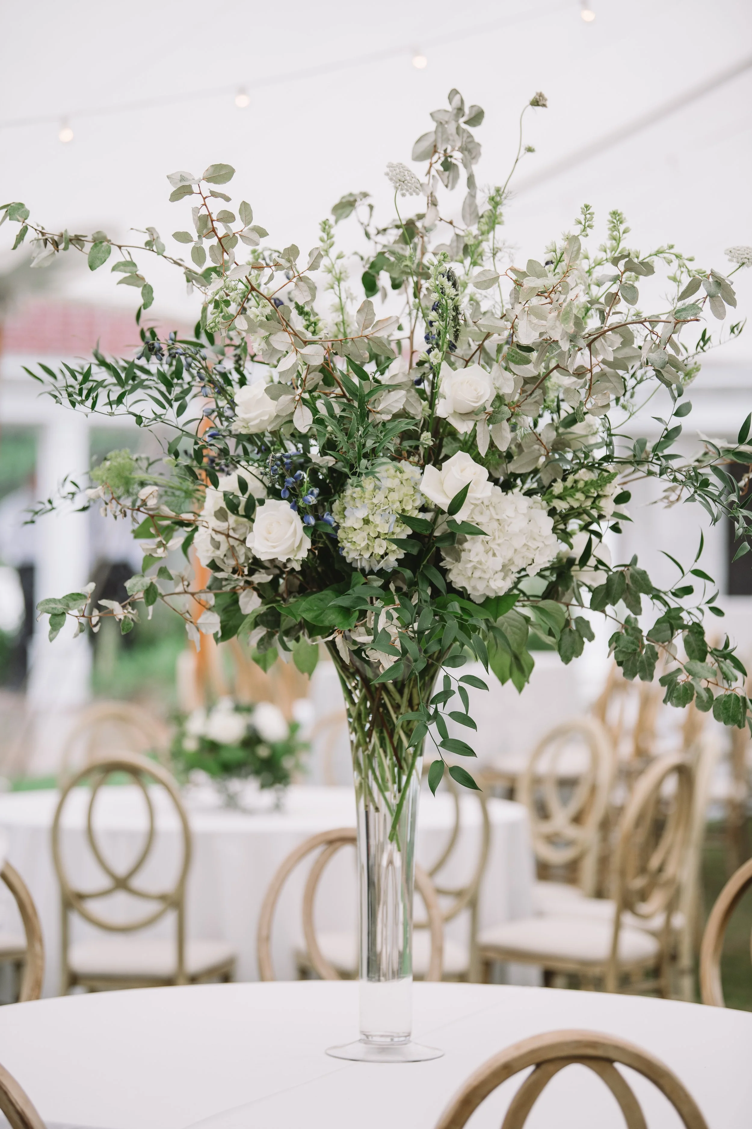 A floral arrangement placed on one of the reception tables. It is in a tall thin vase and includes white roses and other flowers and greenery.