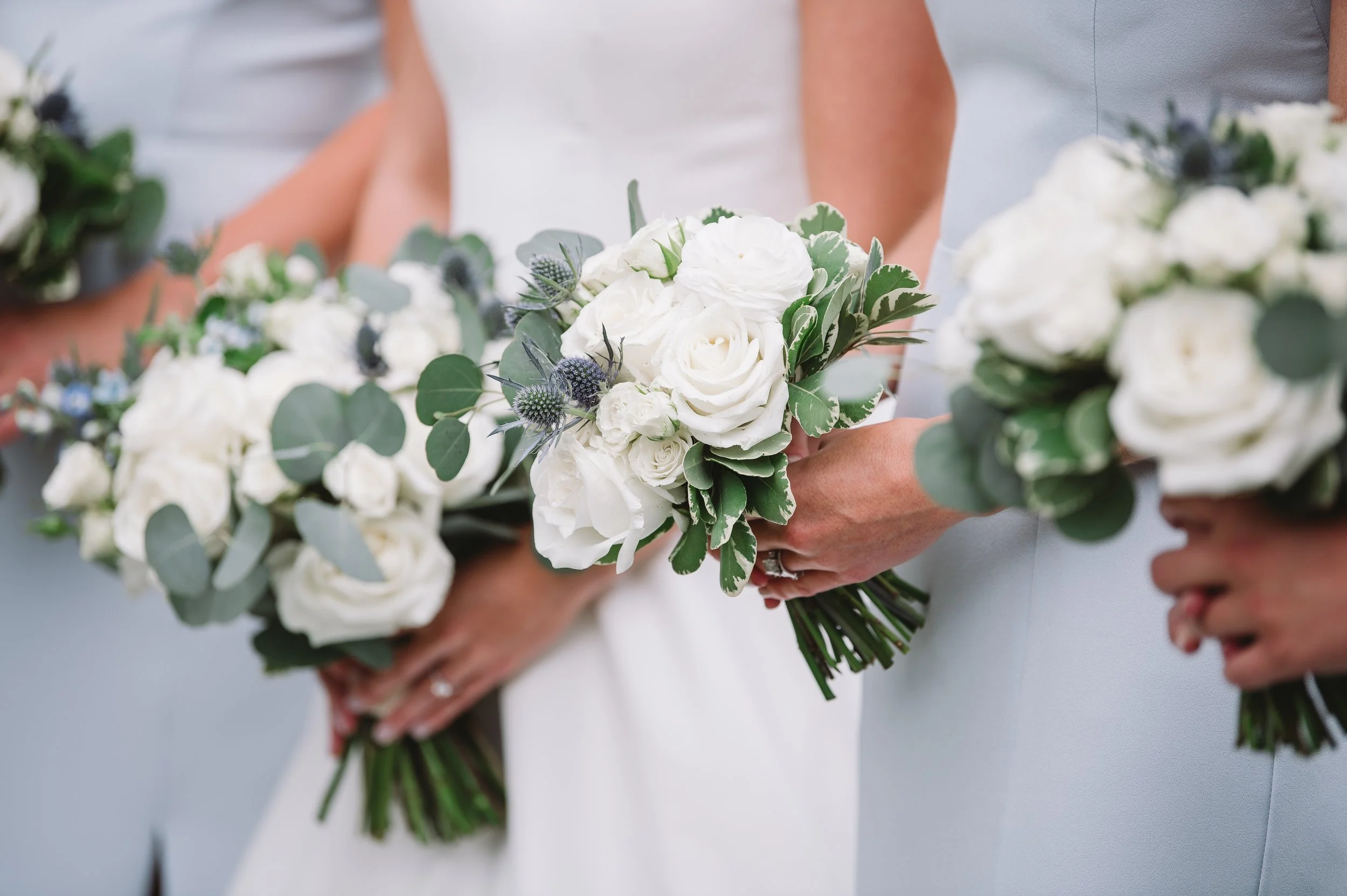 A close up of Callie and her bridesmaids' bouquets. The bouquets have white flowers including roses and greenery. Callie's bouquet has blue flowers as well.