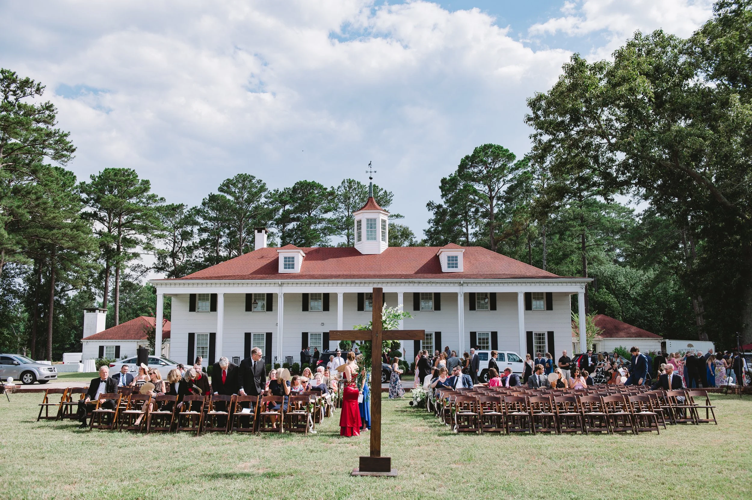 A wide shot of the ceremony spot. In the background there are trees and a large white house with a red roof. Wooden chairs are set up with a wooden cross and bouquet set up in front.