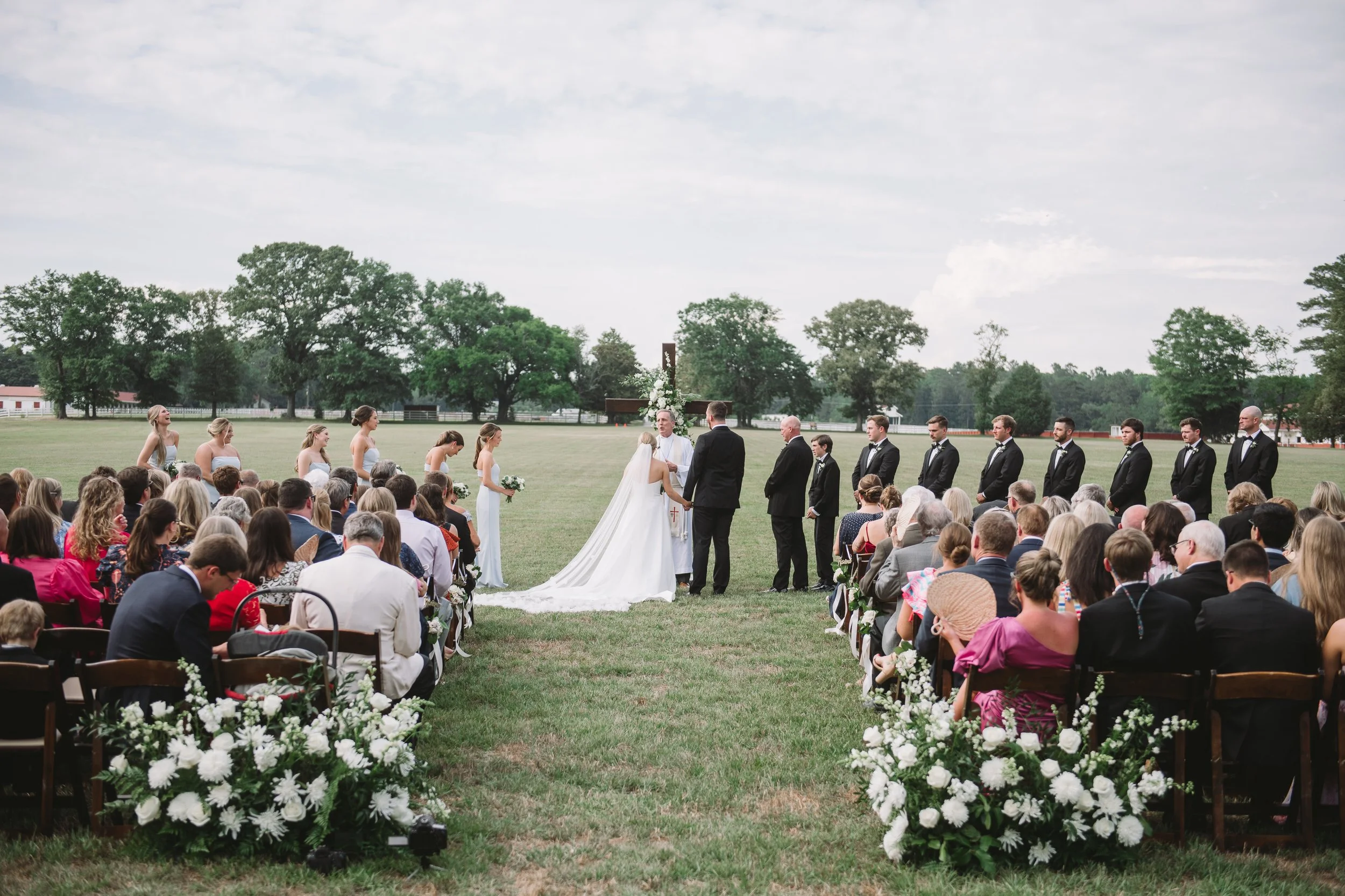 A wide shot taken from the back of the ceremony. Callie and John are in the center with their guests sitting in the foreground. Two large floral arrangements sit behind the last row of chairs.