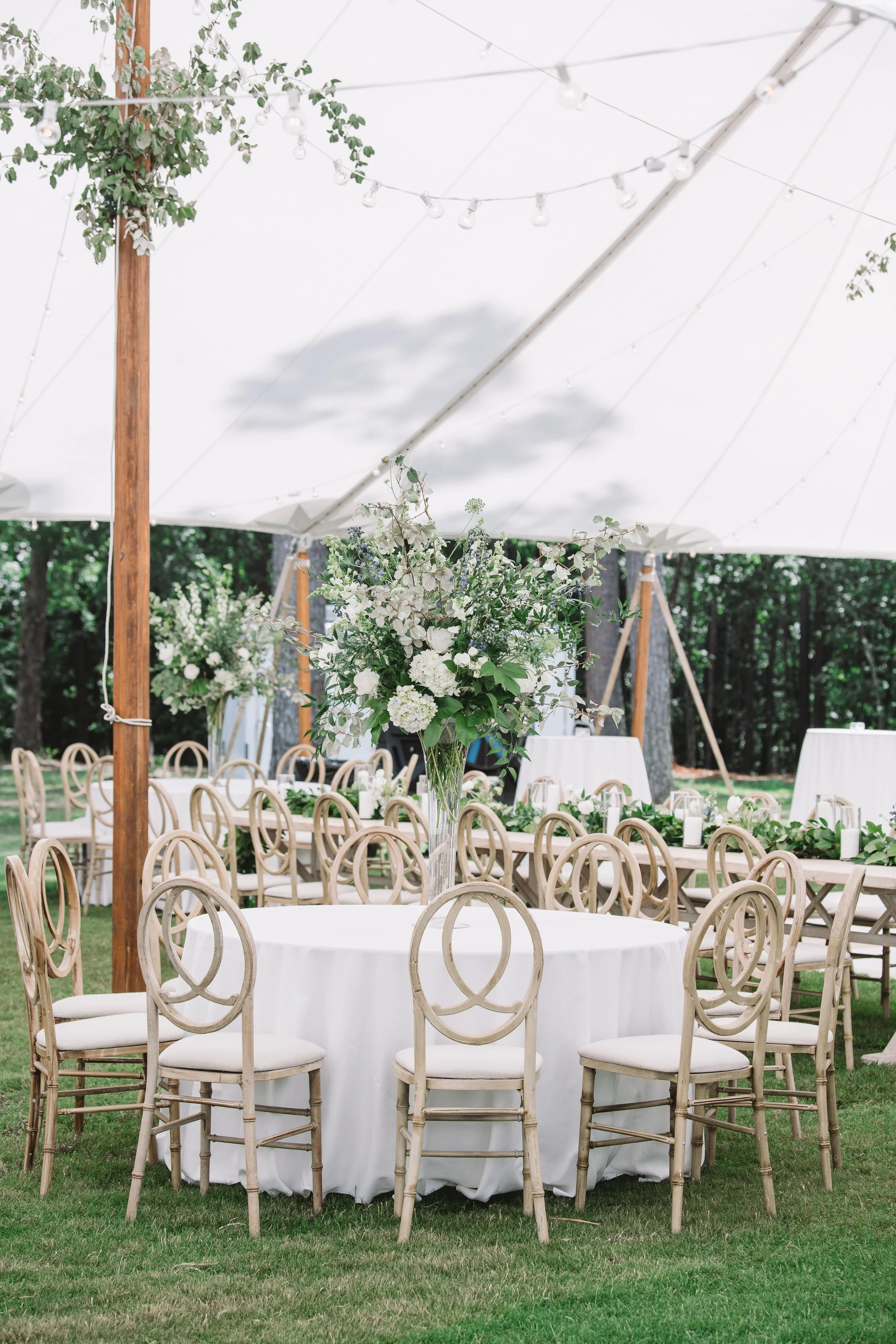 A wide shot of the reception tent. There are tables set up with a showpiece floral arrangement on one of them. The others have small bouquets and candles.