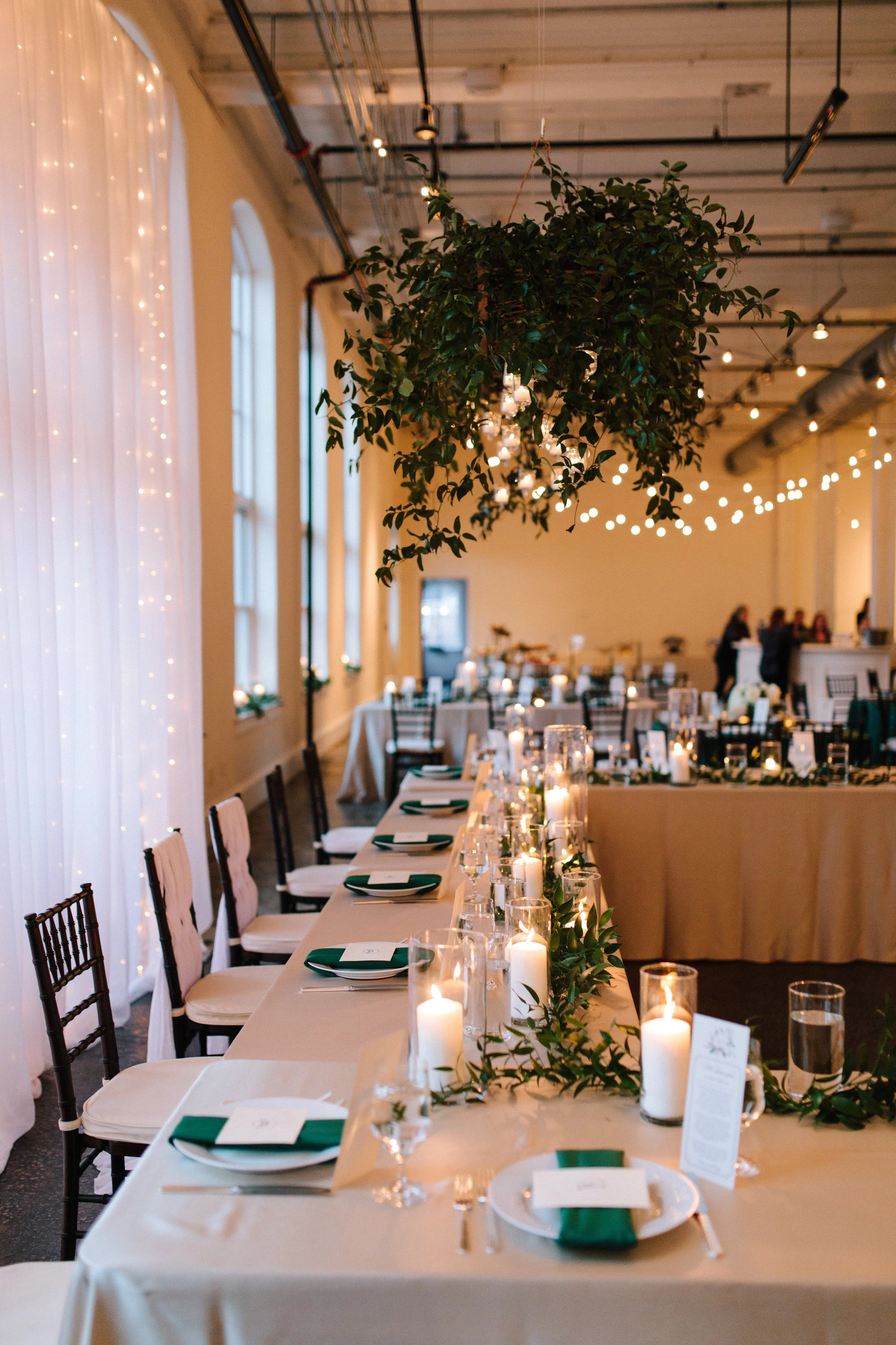 A shot of the reception hall showing a large hanging green vine statement piece and the party's table. There are place settings and white candles and vines as decor.