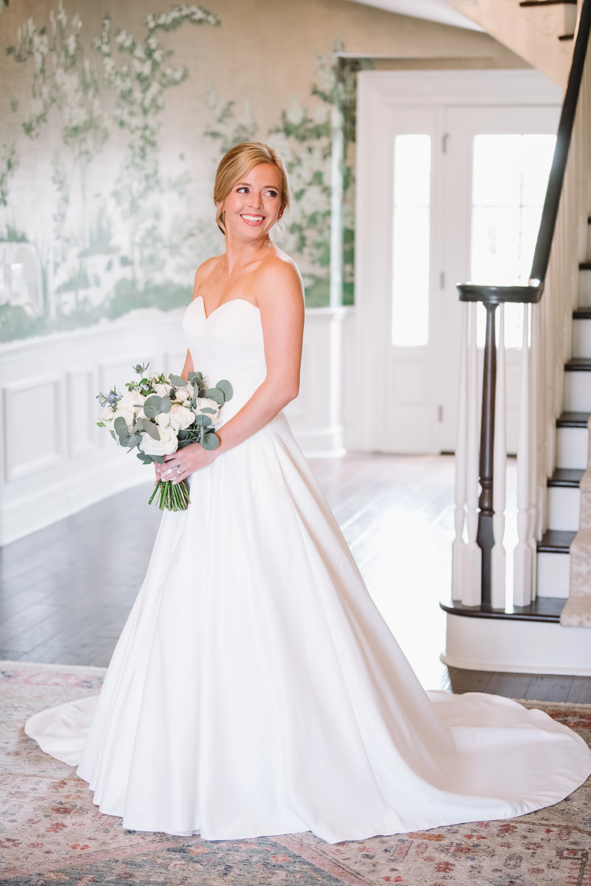 A portrait of Callie looking over her shoulder. She is inside the house and is holding her bouquet. The bouquet has blue and white flowers and greenery.