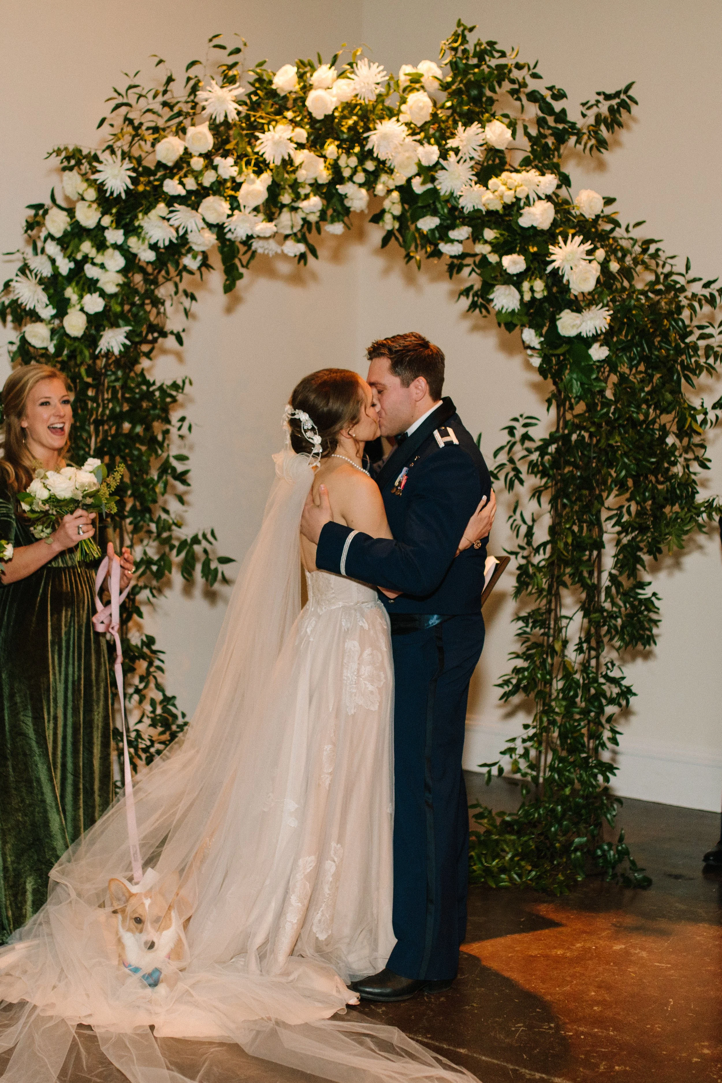 Emily and Hunter kiss at the end of the ceremony underneath a flower arch. The arch is made up of white flowers including roses and green vines.