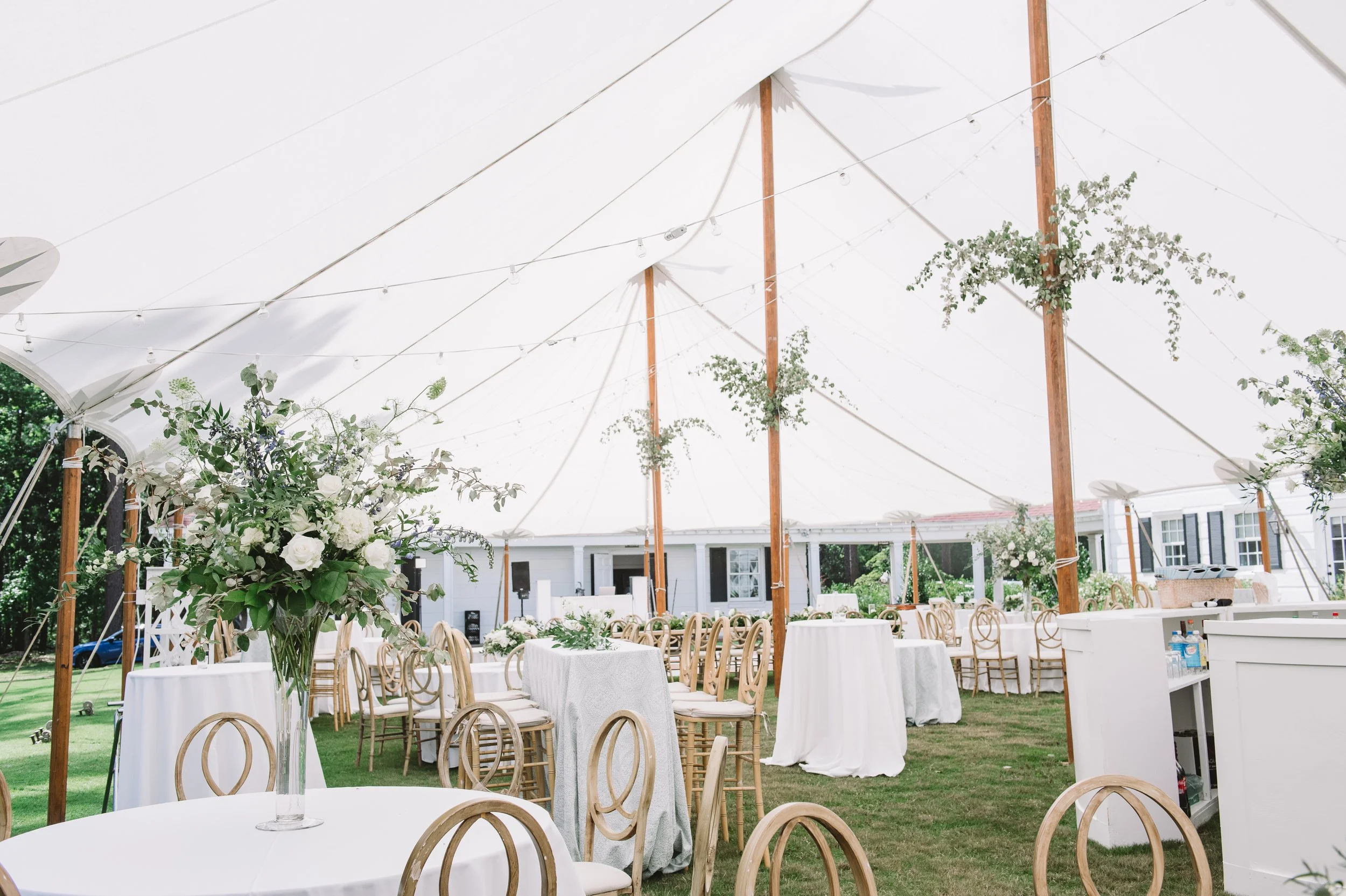 A wide shot of the reception tent. There are tables set up with a showpiece floral arrangement on one of them. The others have small bouquets and candles.