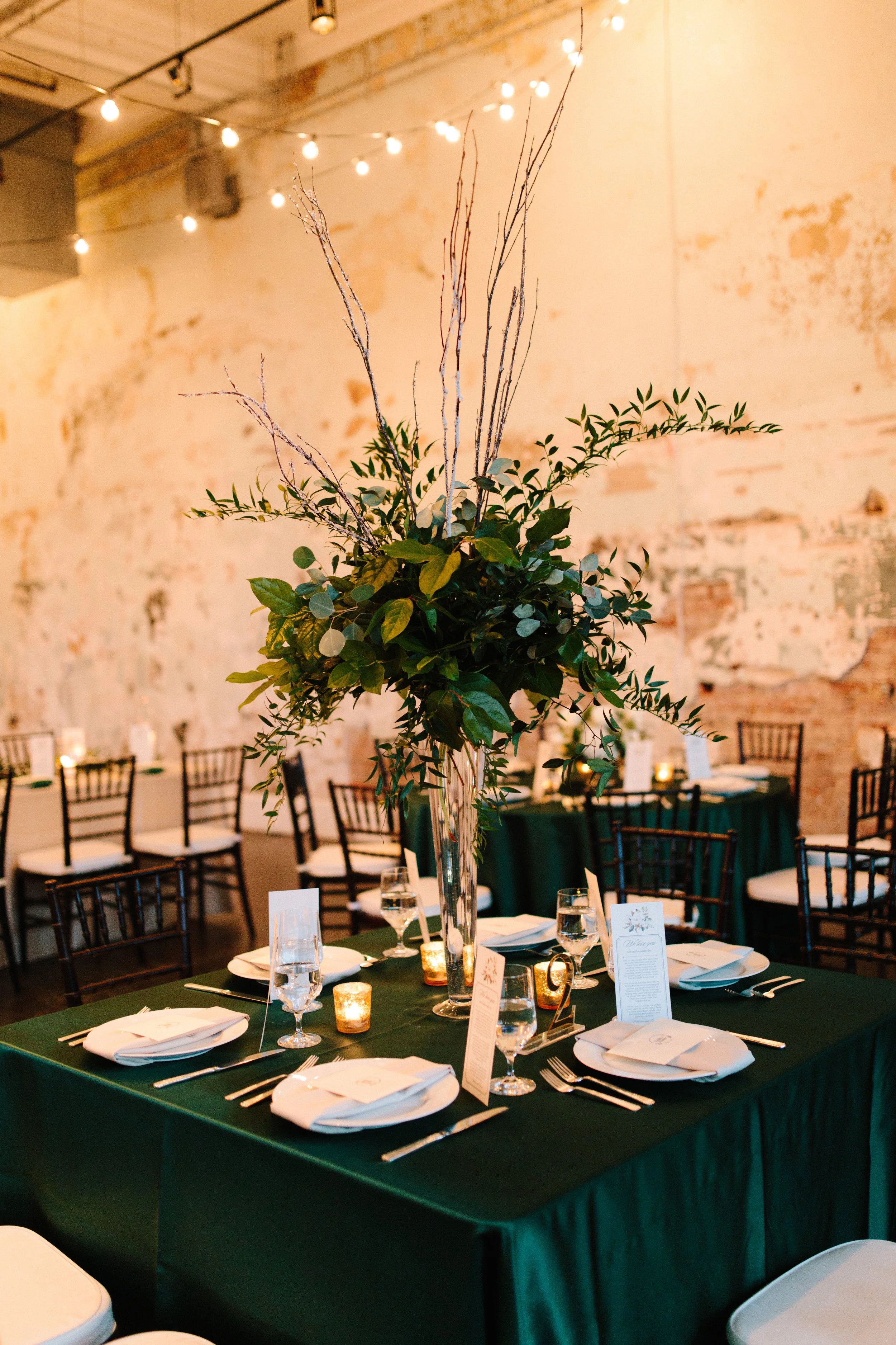 A large flower arrangement set up on one of the dinner tables. The arrangement is in a tall thin vase and is overflowing with greenery. The table has a dark green tablecloth and place settings.