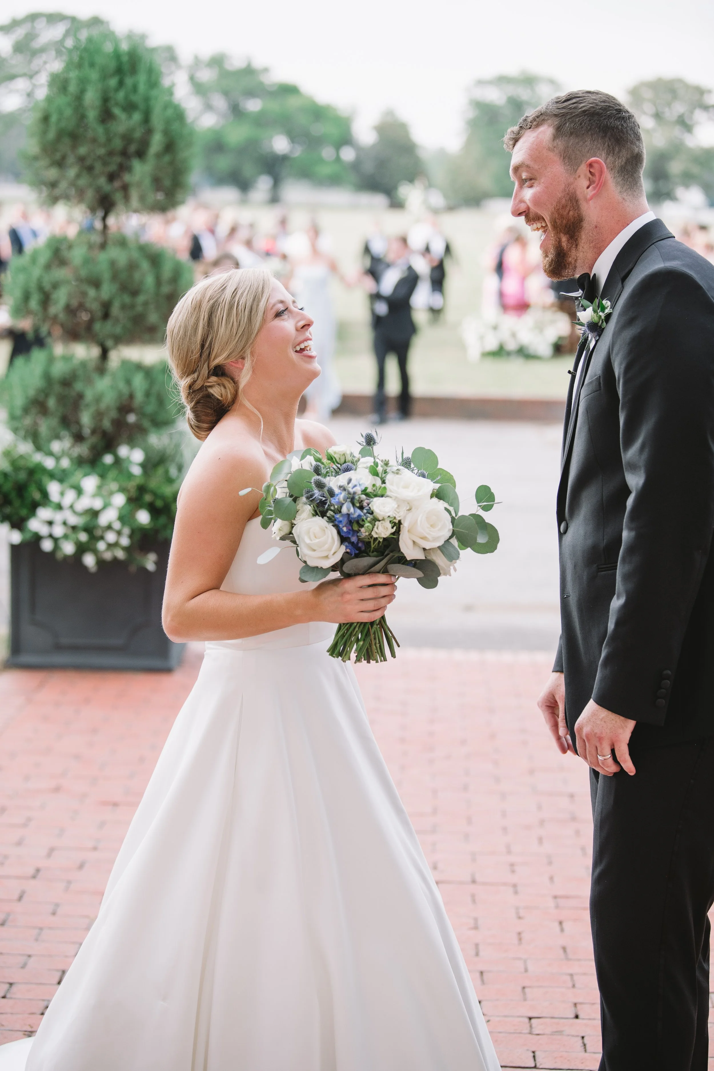 Callie and John laughing with each other. Callie is holding her bouquet which contains white and blue flowers with greenery.