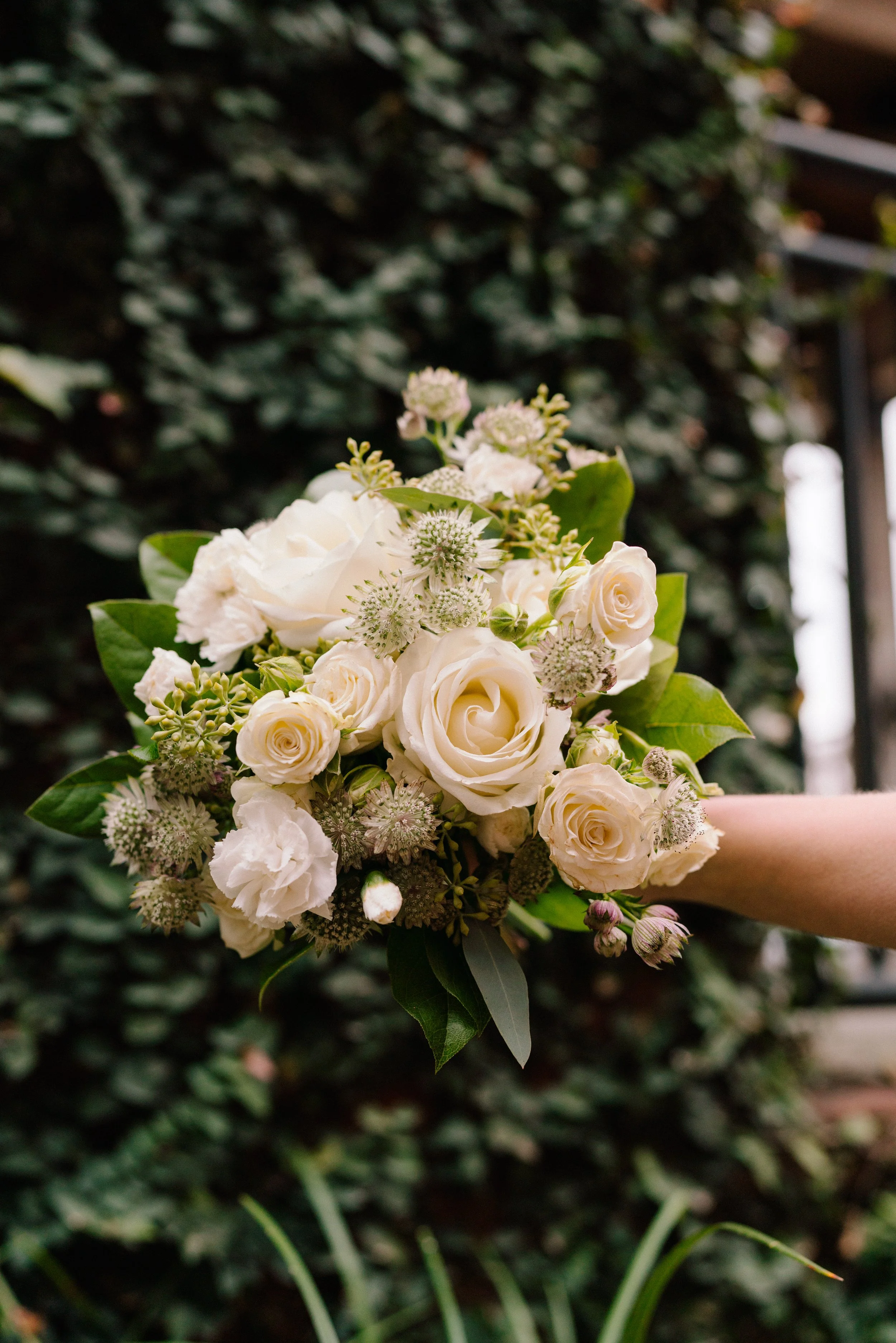 A close up shot of a bouquet containing white flowers and leaves. There is an ivy-covered wall in the background.
