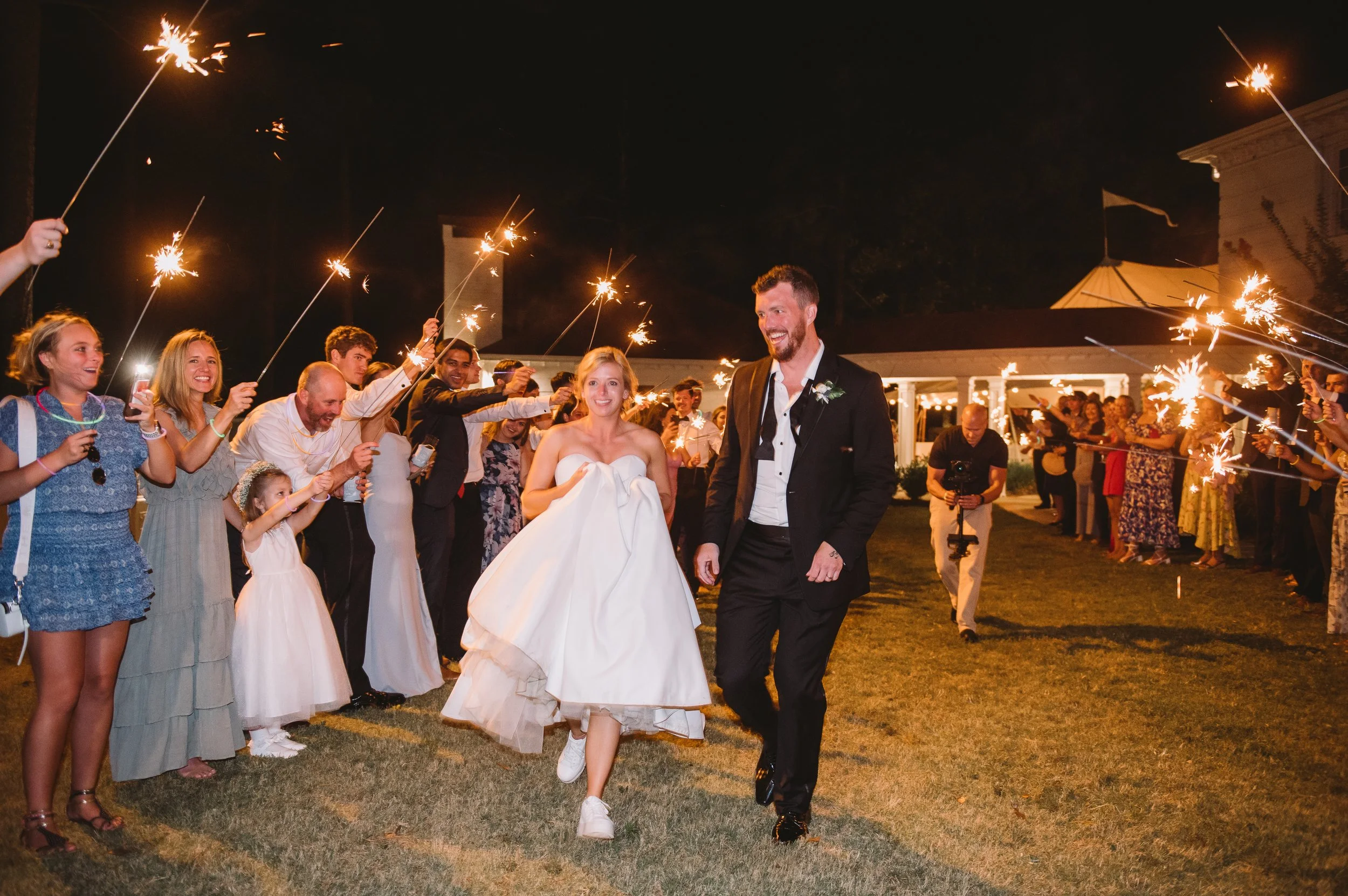 Callie and John leaving their reception with a sparkler send off. Both are smiling and running through the guests who are holding sparklers above their heads.