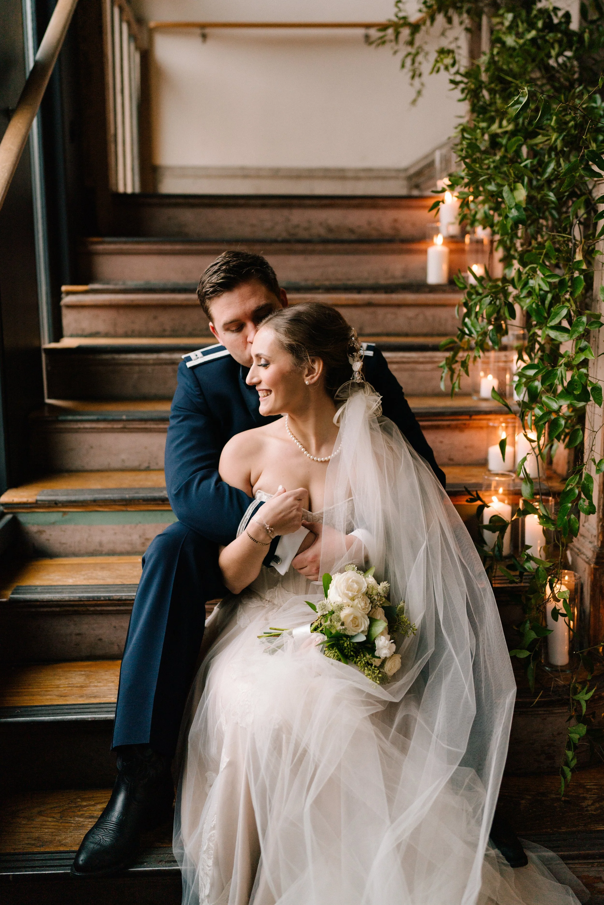 Emily and Hunter sit on a wooden stair case, she smiles as he kisses her cheek. There are trailing vines and candles on the side of the stairs.