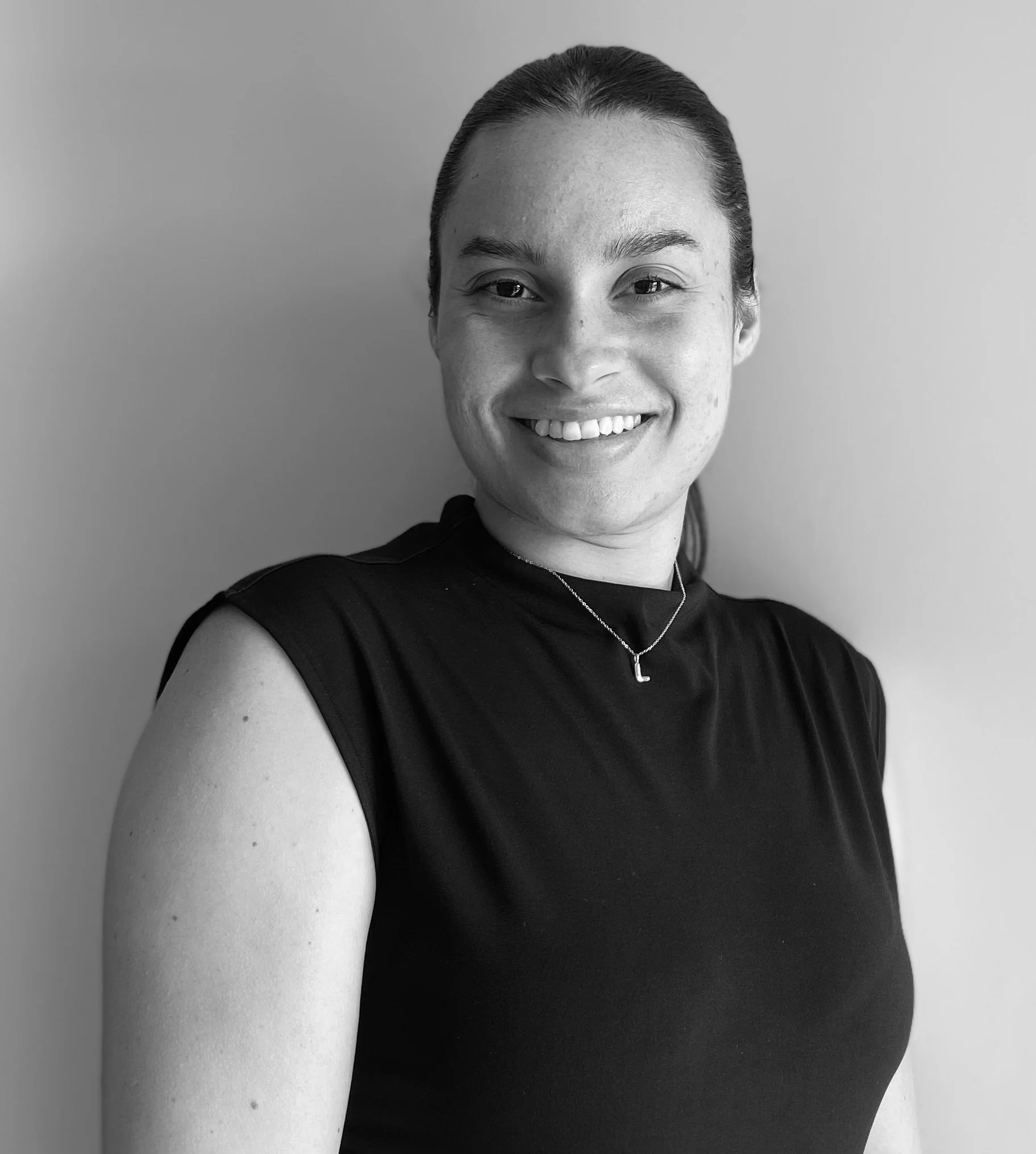 Black and white portrait of a smiling woman with her hair tied back, wearing a sleeveless top and a necklace with a letter 'L', standing against a plain background.
