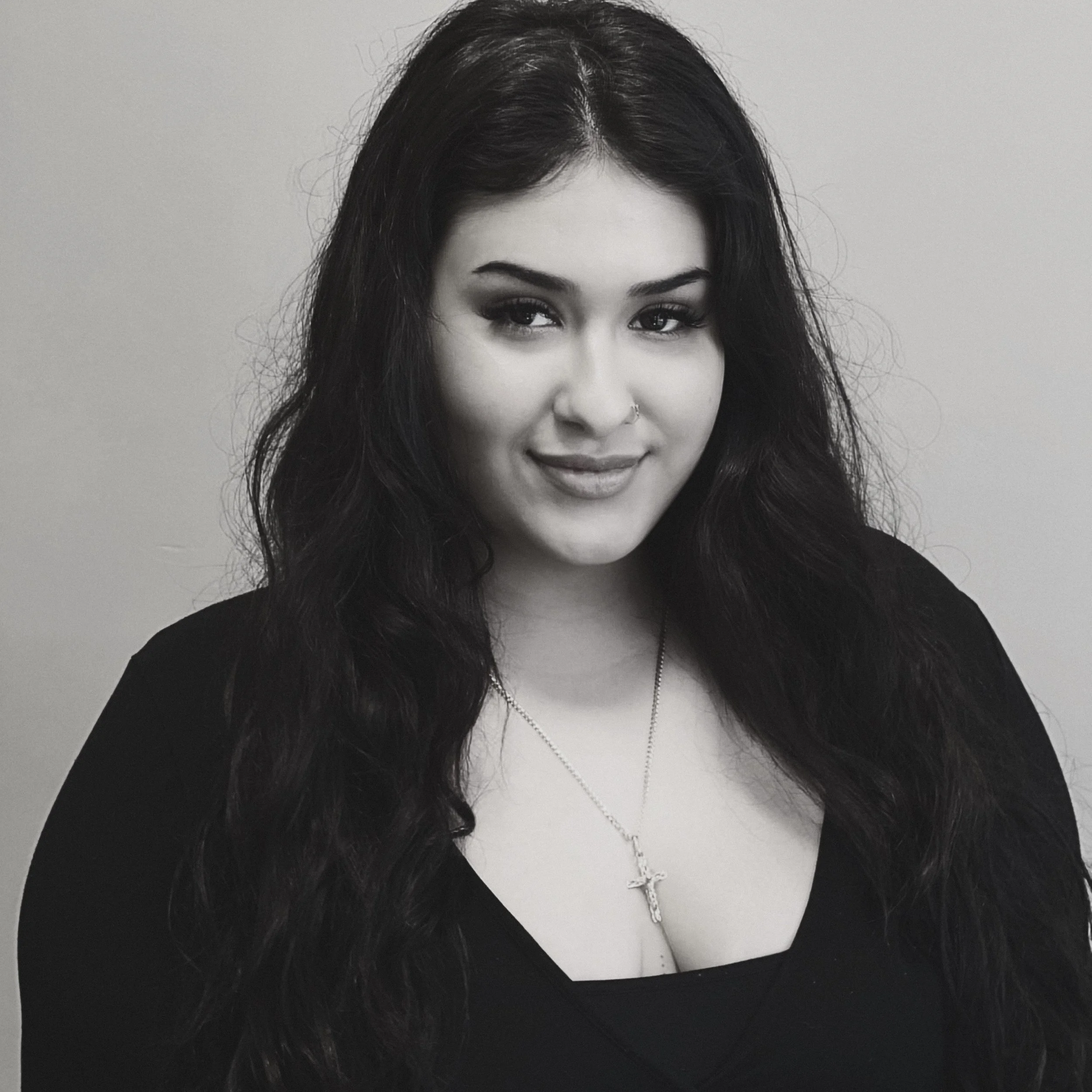 Black and white portrait of a smiling young woman with long dark hair, wearing a necklace with a cross pendant and a black top, against a plain background.