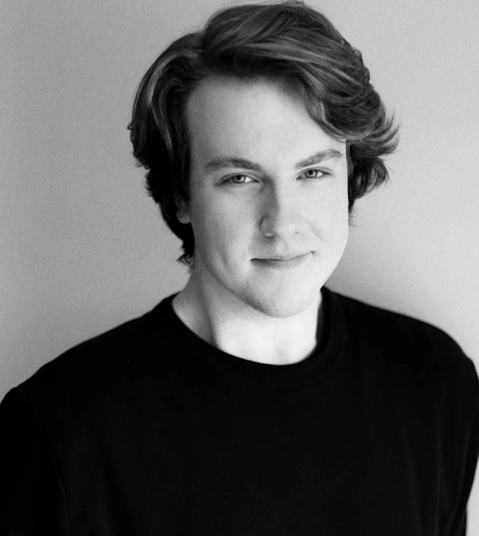 Black and white portrait of a young person with wavy hair wearing a black shirt, looking slightly to the side with a subtle smile.