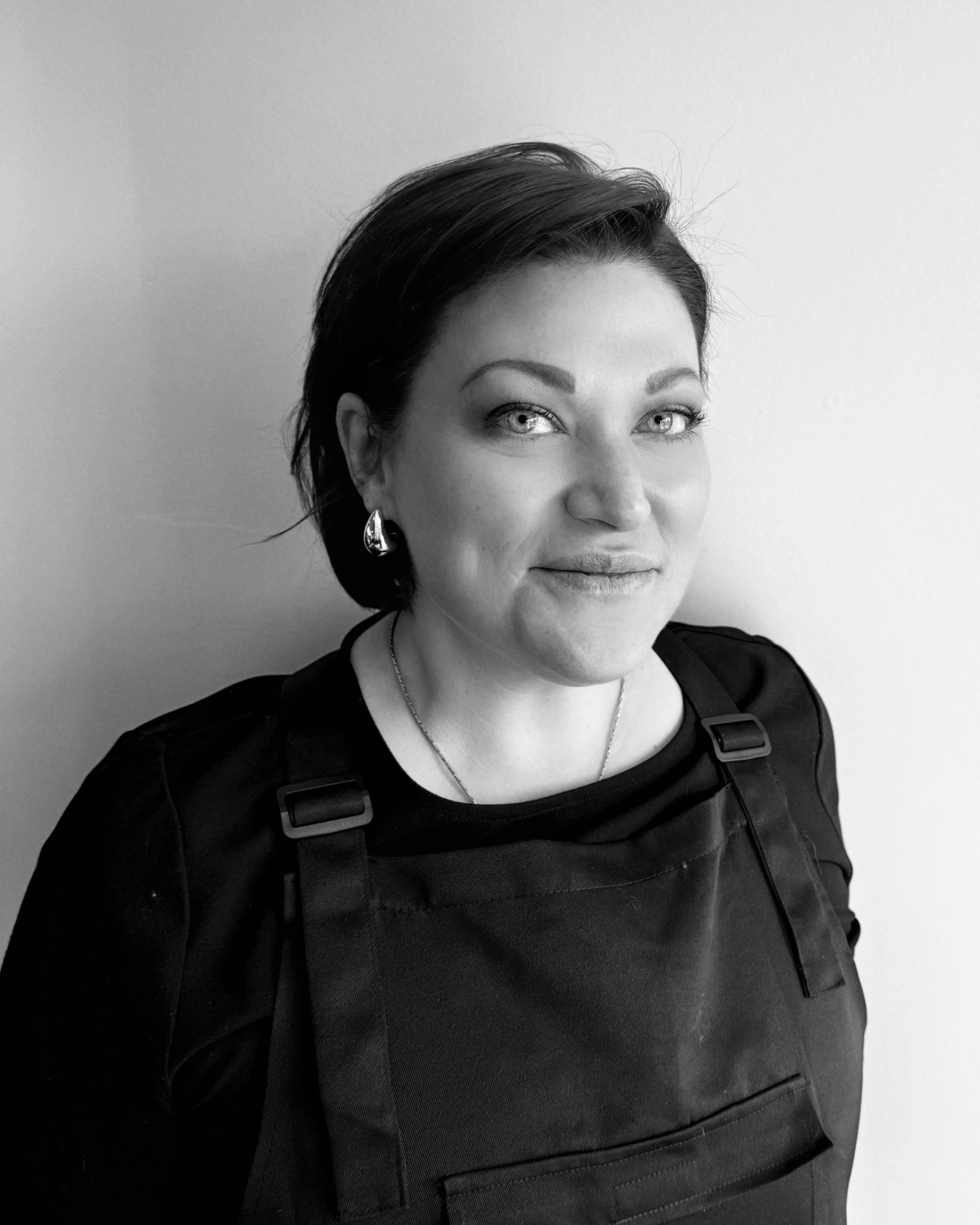 Black and white photo of a woman with short dark hair, earrings, and a black top, smiling slightly, standing against a plain light-colored wall.