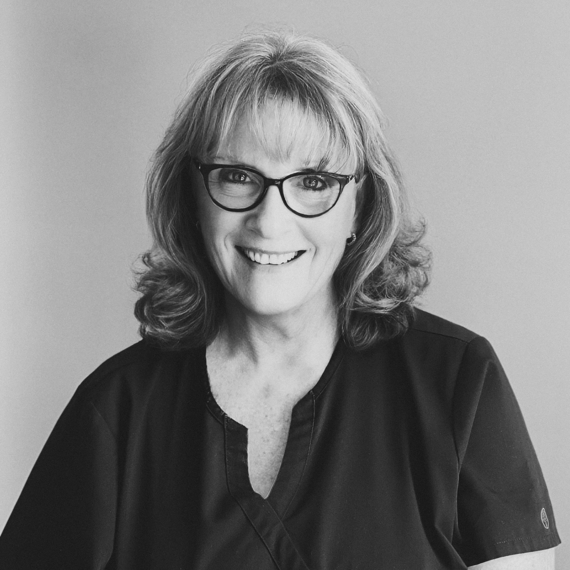 Black and white portrait of a smiling woman with glasses, shoulder-length wavy hair, wearing a dark scrub top, against a plain light-colored background.