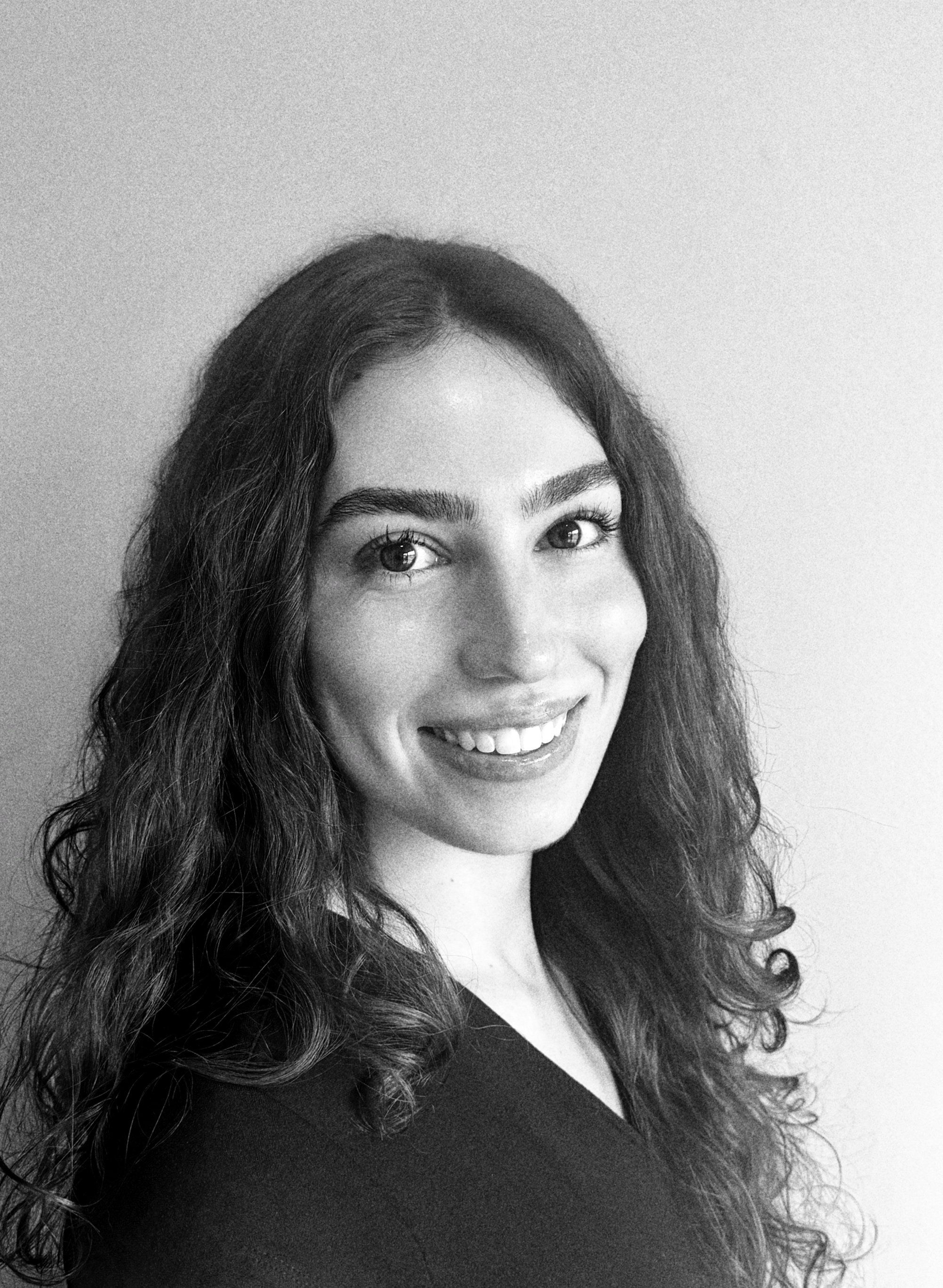 Black and white portrait of a smiling woman with wavy hair, wearing a dark top and standing against a plain light-colored background.
