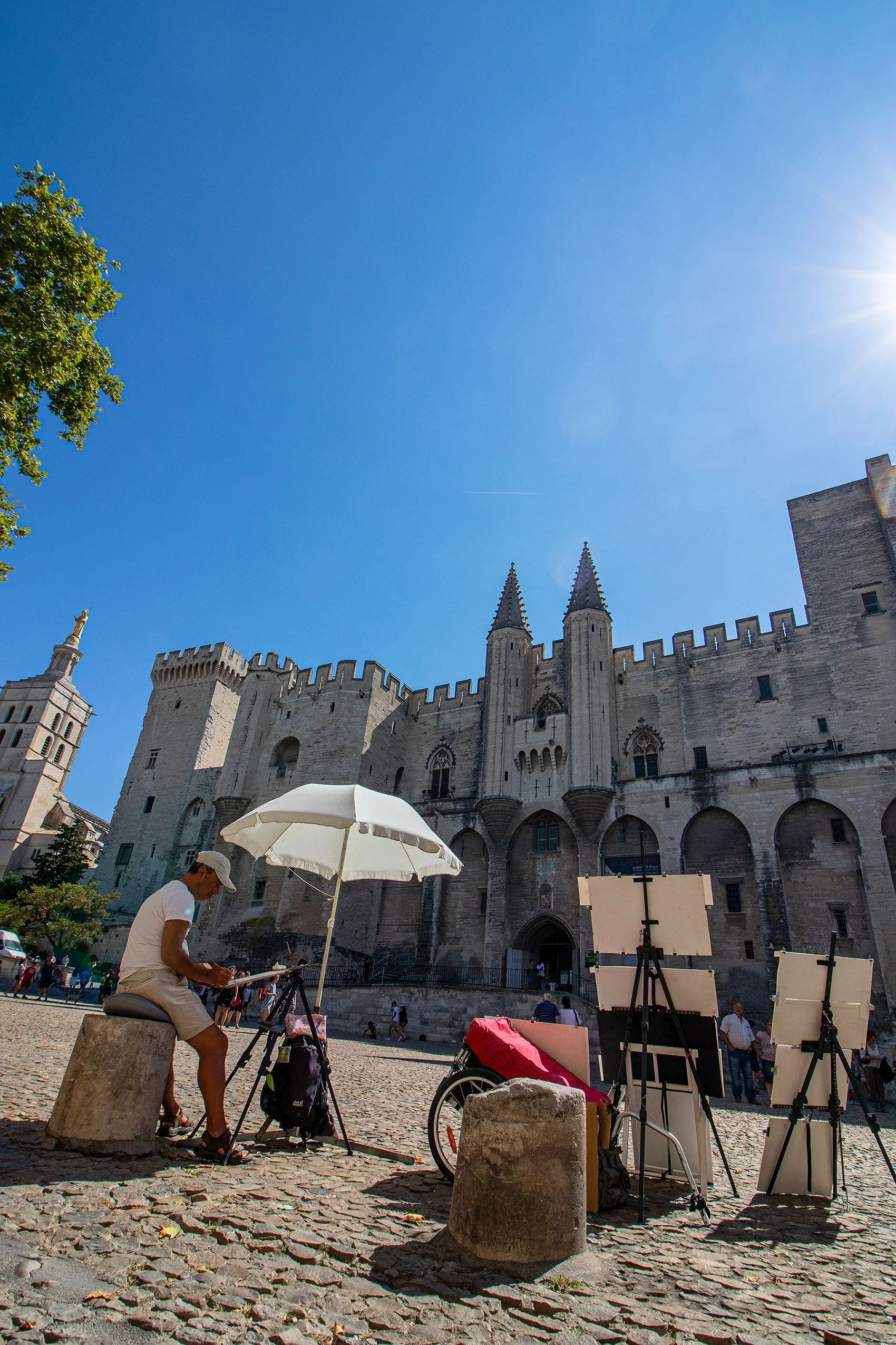 Beeld van een man die op een bankje zit in Avignon met een parasol, omringd door schildersezels, met een middeleeuws kasteel op de achtergrond onder een heldere blauwe hemel.