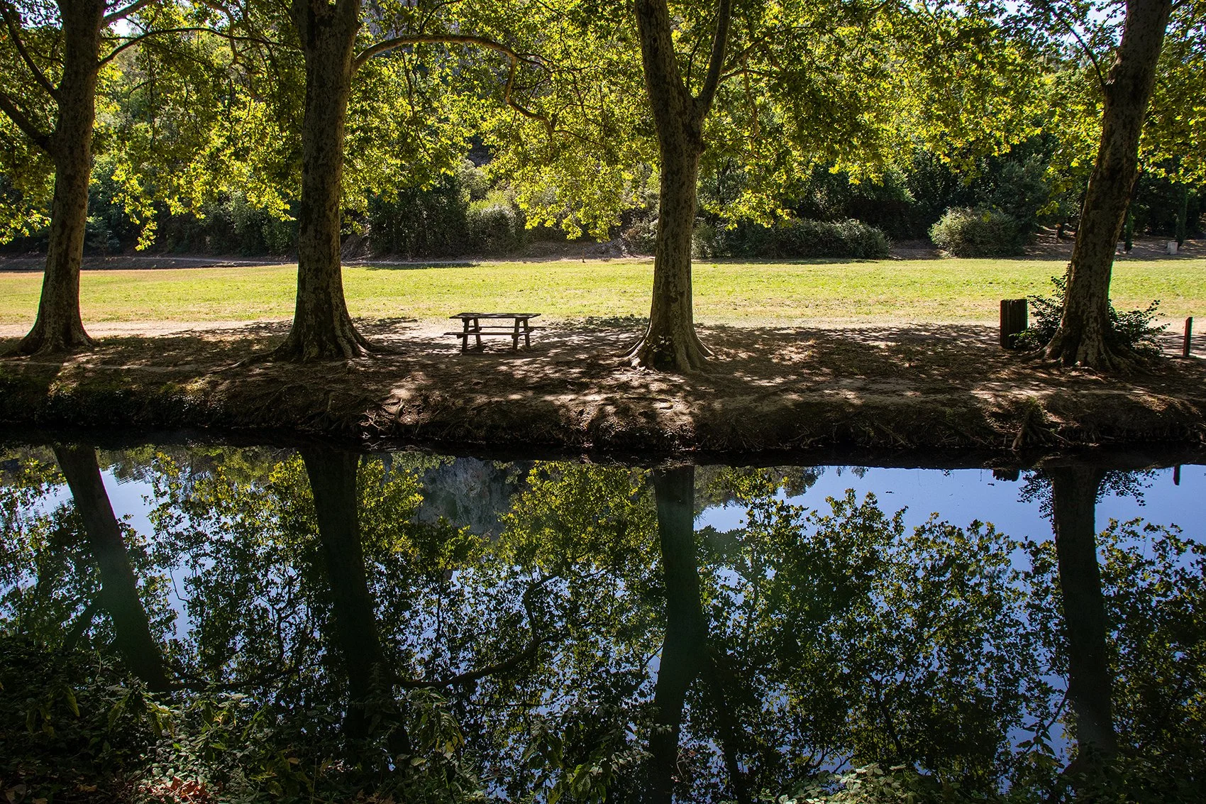 Een park met drie grote bomen, bij de rivier de Eure, een bankje onder de middelste boom, en een vijver die de reflection van de bomen en de blauwe lucht met witte wolken weergeeft.
