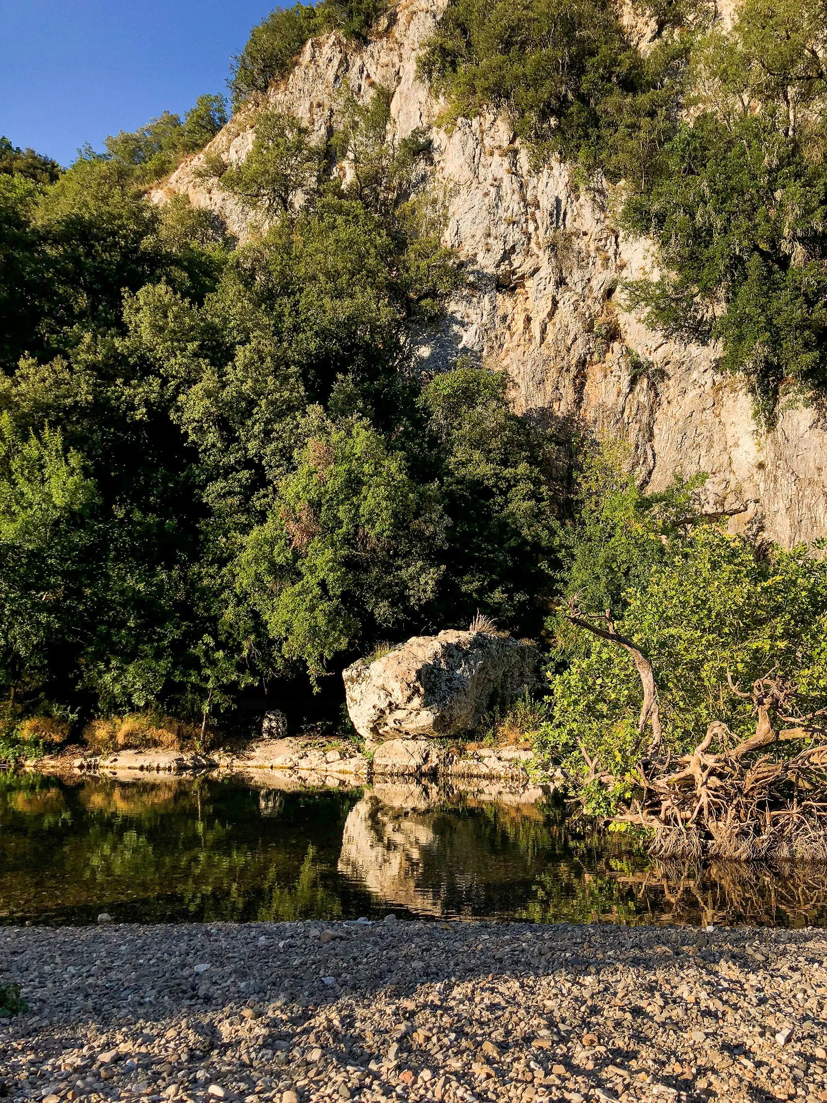 Natuursteen met bomen en rotsen naast een rustige rivier de Cèze, weerspiegeling in het water, onder een blauwe hemel.