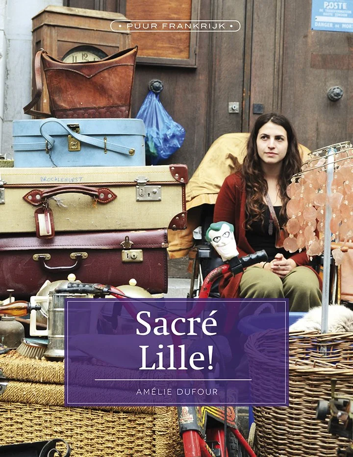 Een vrouw zit tussen geruime vintage spullen, waaronder koffers, tassen, en een fiets, op een antiekmarkt, met een bordje met de tekst 'Sacré Lille!' en haar naam Amélie Dufour. Er worden ook Godzilla en andere speelgoedpoppen getoond.