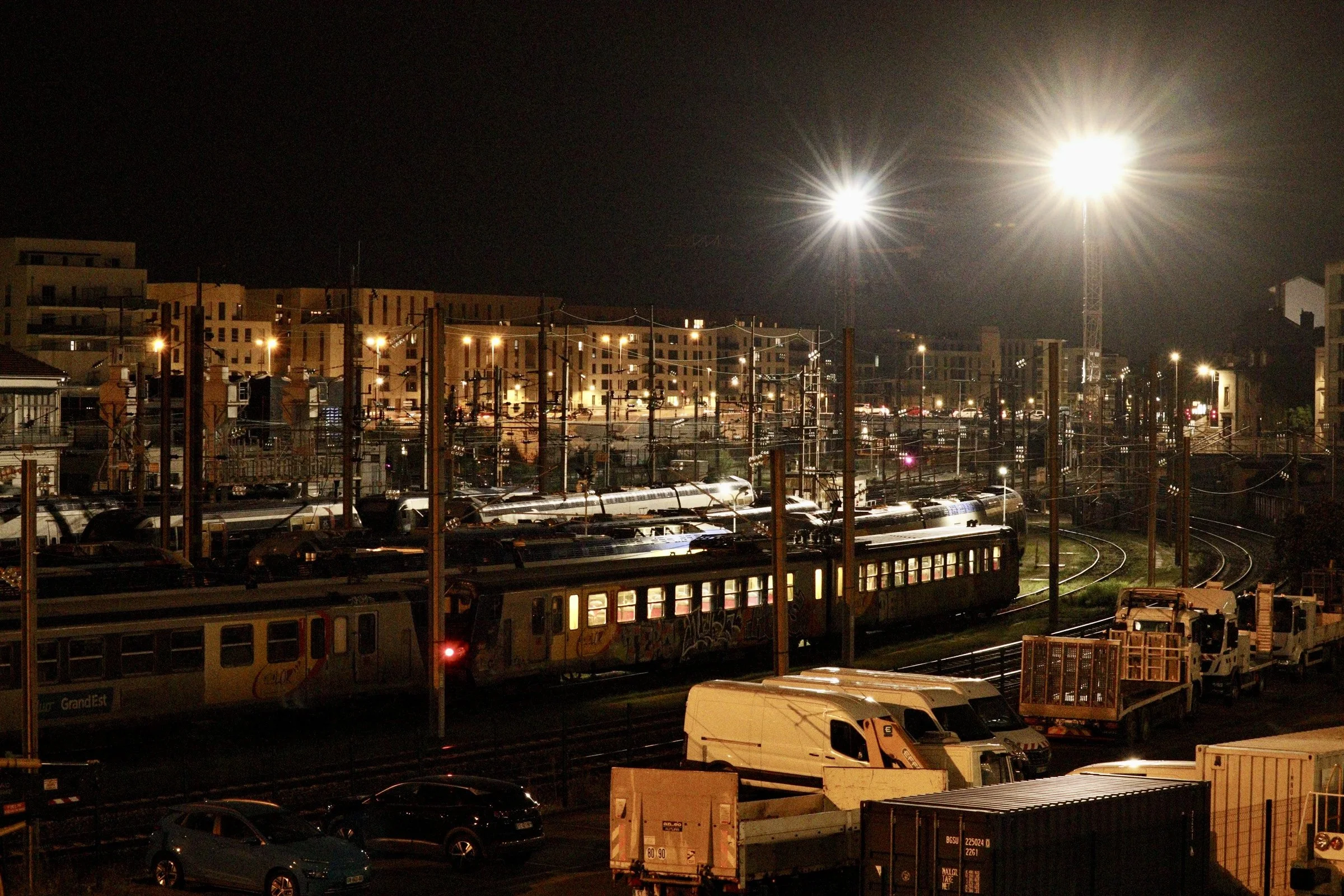 Een trein op het spoor bij nacht met veel lichtbronnen en omringende voertuigen.