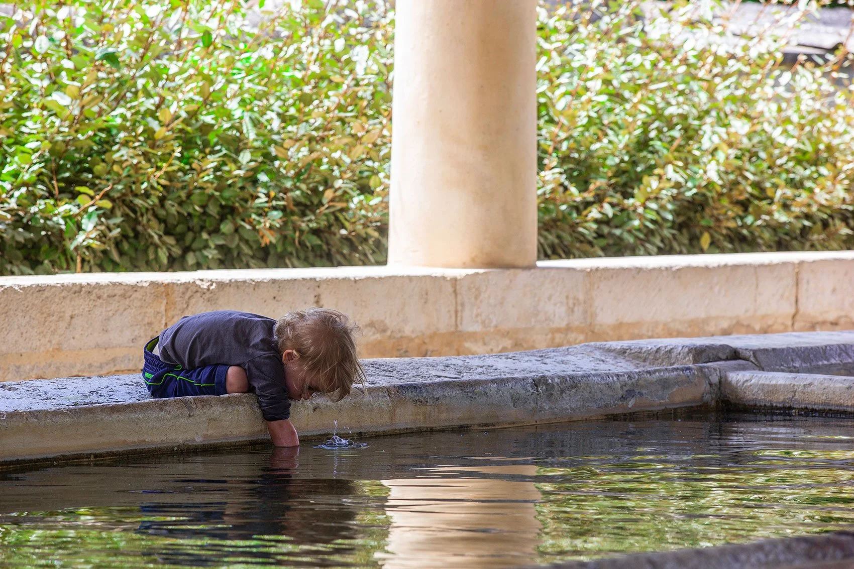 Een jong kind dat met zijn hand in het water van een fontein speelt, onder een boog met groene struiken op de achtergrond.