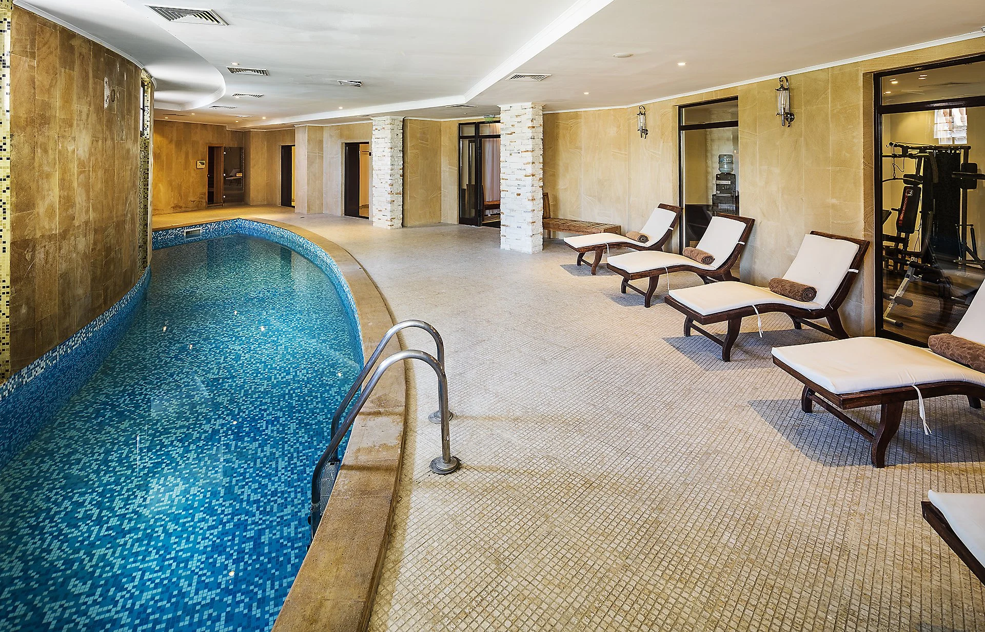 Interior of an indoor swimming pool area with lounge chairs and exercise equipment visible through a glass door.