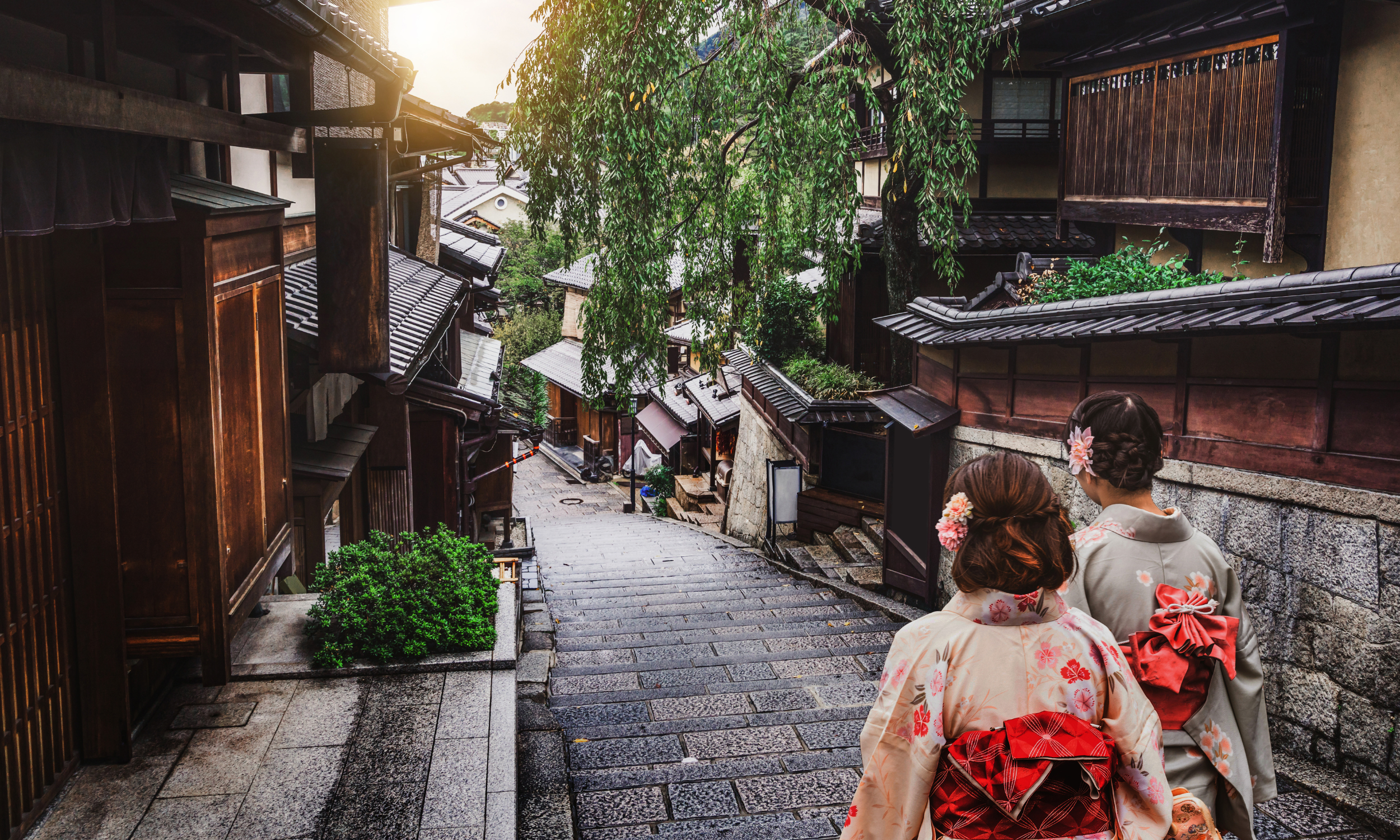 Two women in traditional Japanese kimonos walking down a historic stone-paved street lined with wooden houses and green foliage in Kyoto, Japan.