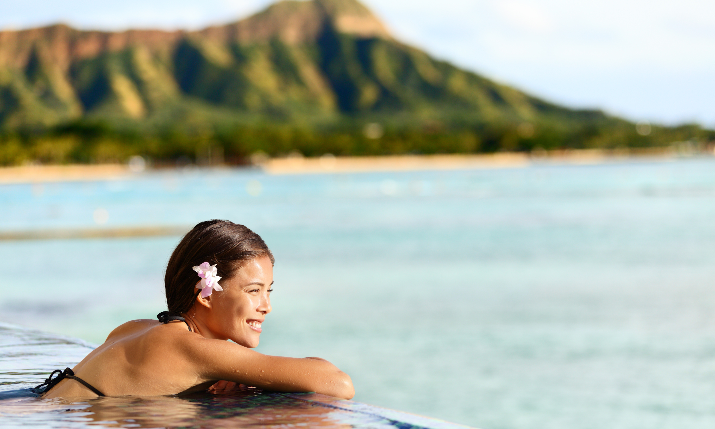 Woman with a flower in her hair relaxing in an infinity pool overlooking a tropical beach with a mountain backdrop