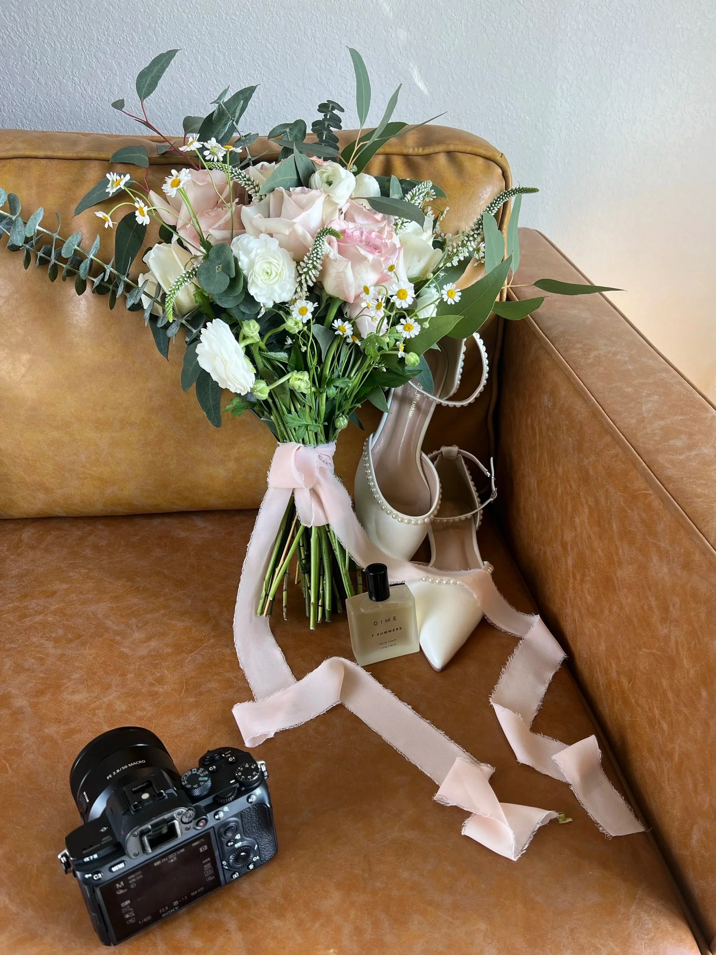 Styled wedding flat lay shot with a pink, white, and green bridal bouquet, pearl-trimmed heels, perfume, and camera displayed on a vintage leather chair—perfect for timeless florist portfolios.