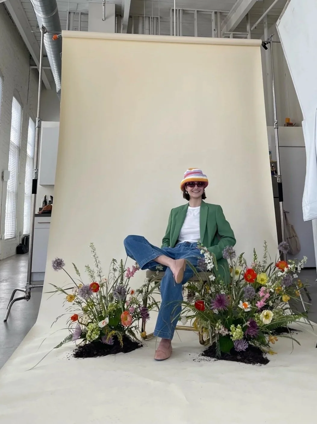 Female floral designer seated within organic, vibrant arrangements mounted in soil on a draped floor, creating a nature-meets-studio aesthetic for a standout brand photoshoot.