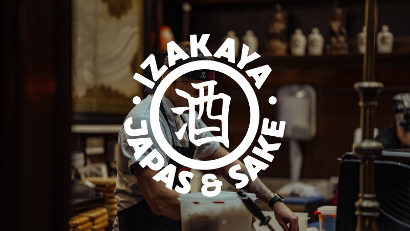 Interior of a Japanese izakaya with a focus on a chef preparing food behind the counter. The overlay text reads 'Izakaya Japas & Sake' with a Japanese character in the middle.