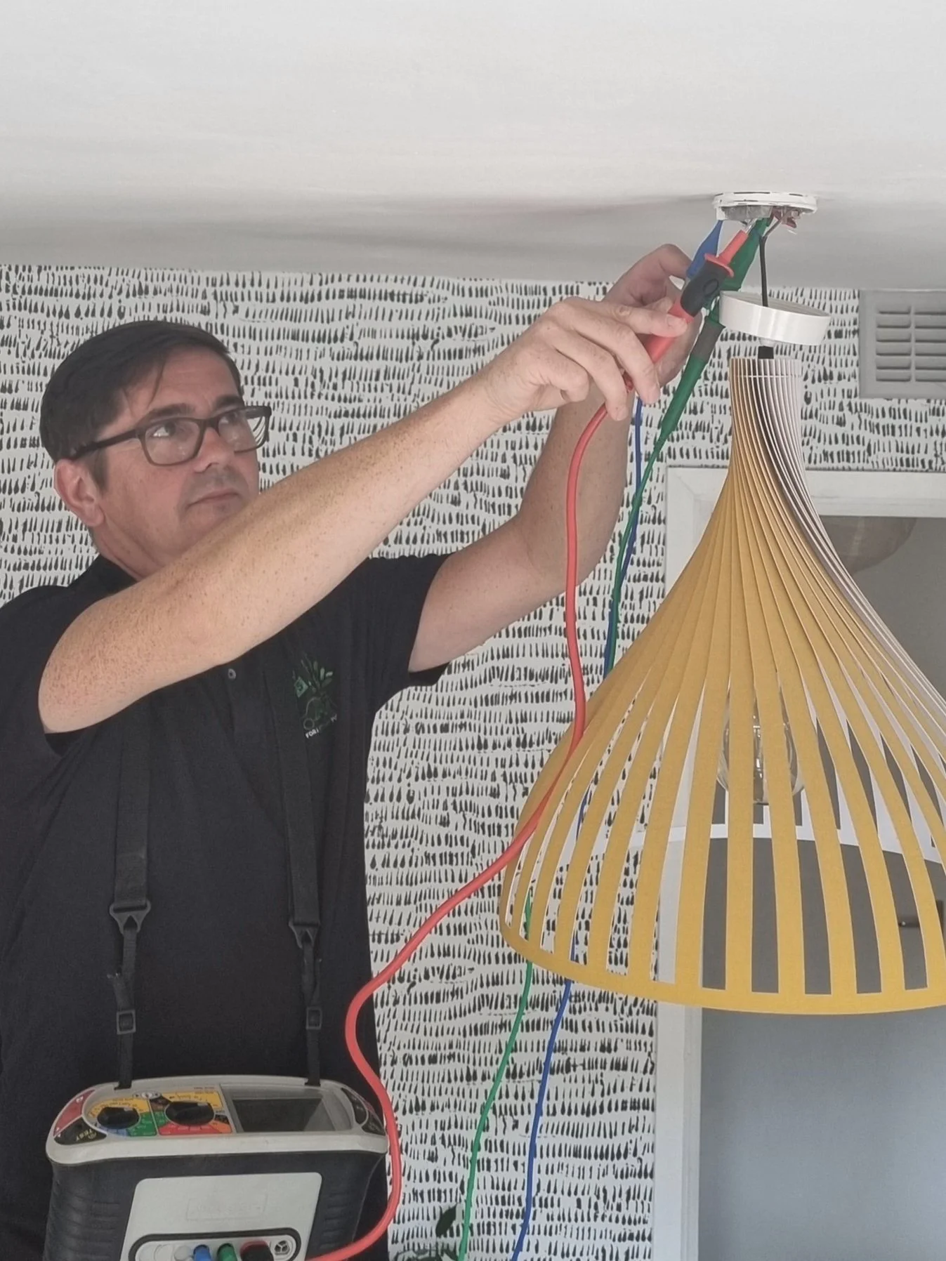 A man wearing glasses and a black shirt is working on a ceiling light fixture, using tools to make electrical connections with red, green, and blue wires.