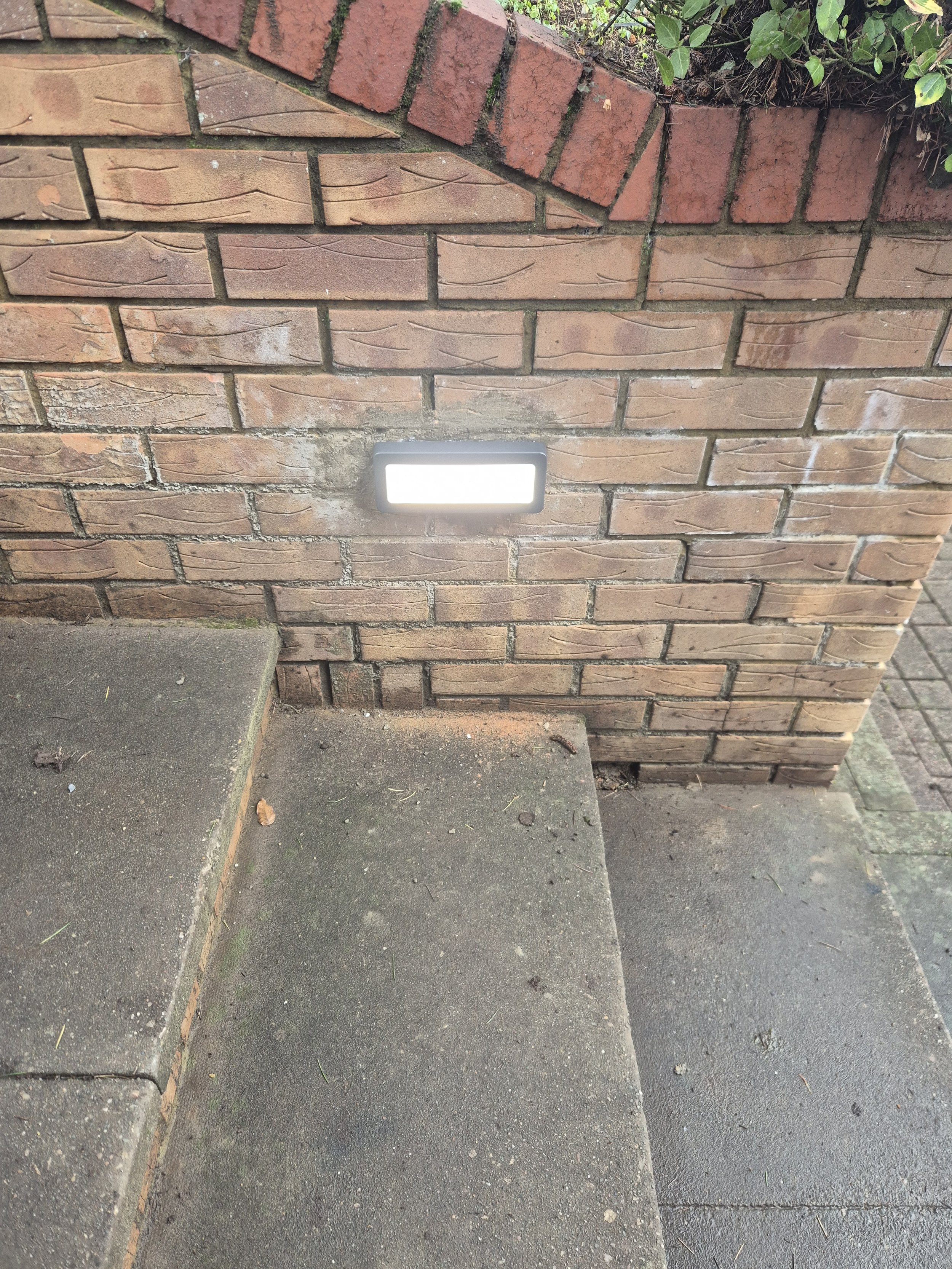 Brick wall with a built-in LED light illuminating the ground on a sidewalk, with concrete steps and some green plants at the top of the wall.