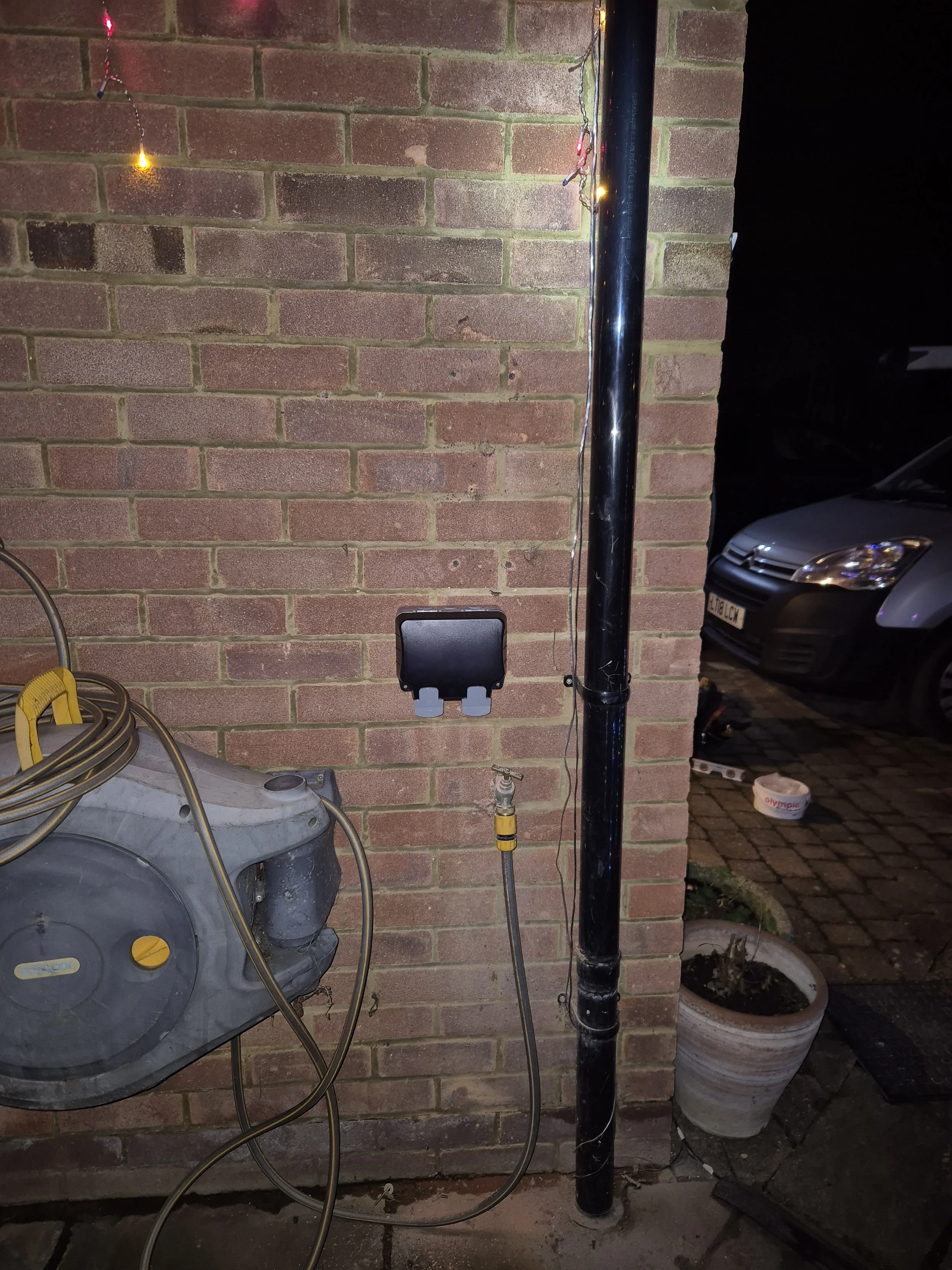 Outdoor scene showing a brick wall with a black pole, a black electrical box, a coiled hose reel, and a potted plant, with a dark car parked on the street.