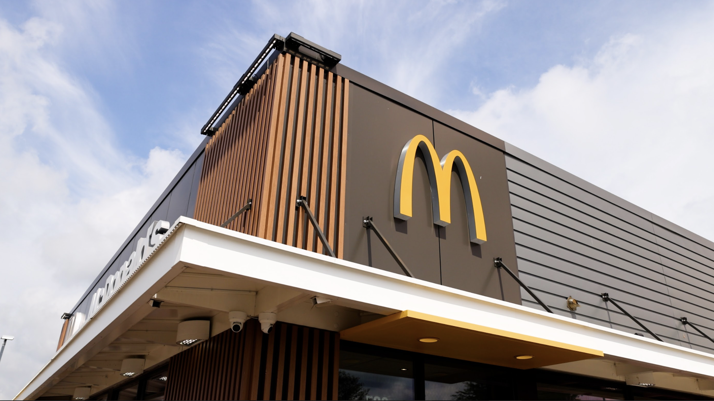 Exterior of a McDonald's restaurant with yellow 'M' logo, wood and gray siding, under a partly cloudy sky.