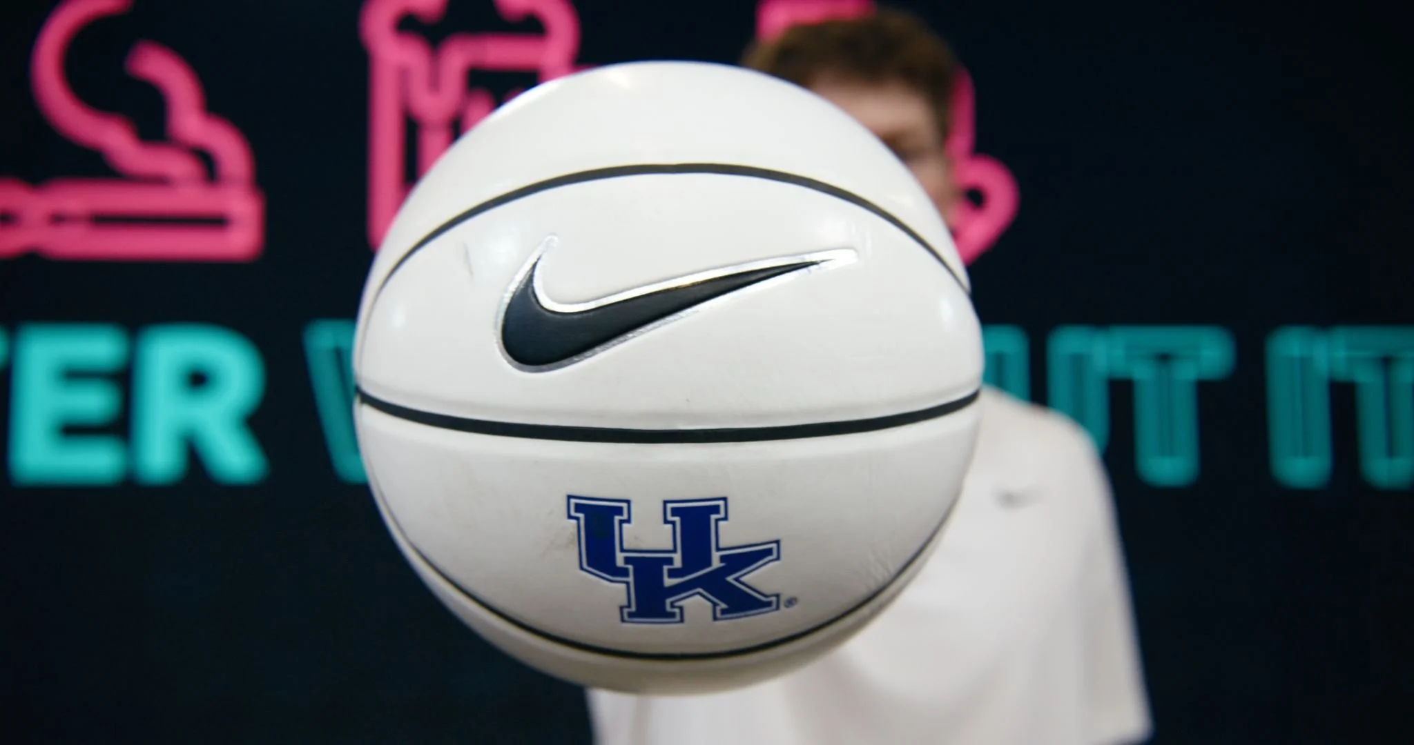 A person holding a white Nike basketball with the Kentucky Wildcats logo in front of their face. The background appears to be a sports or event setting with signs and lights.