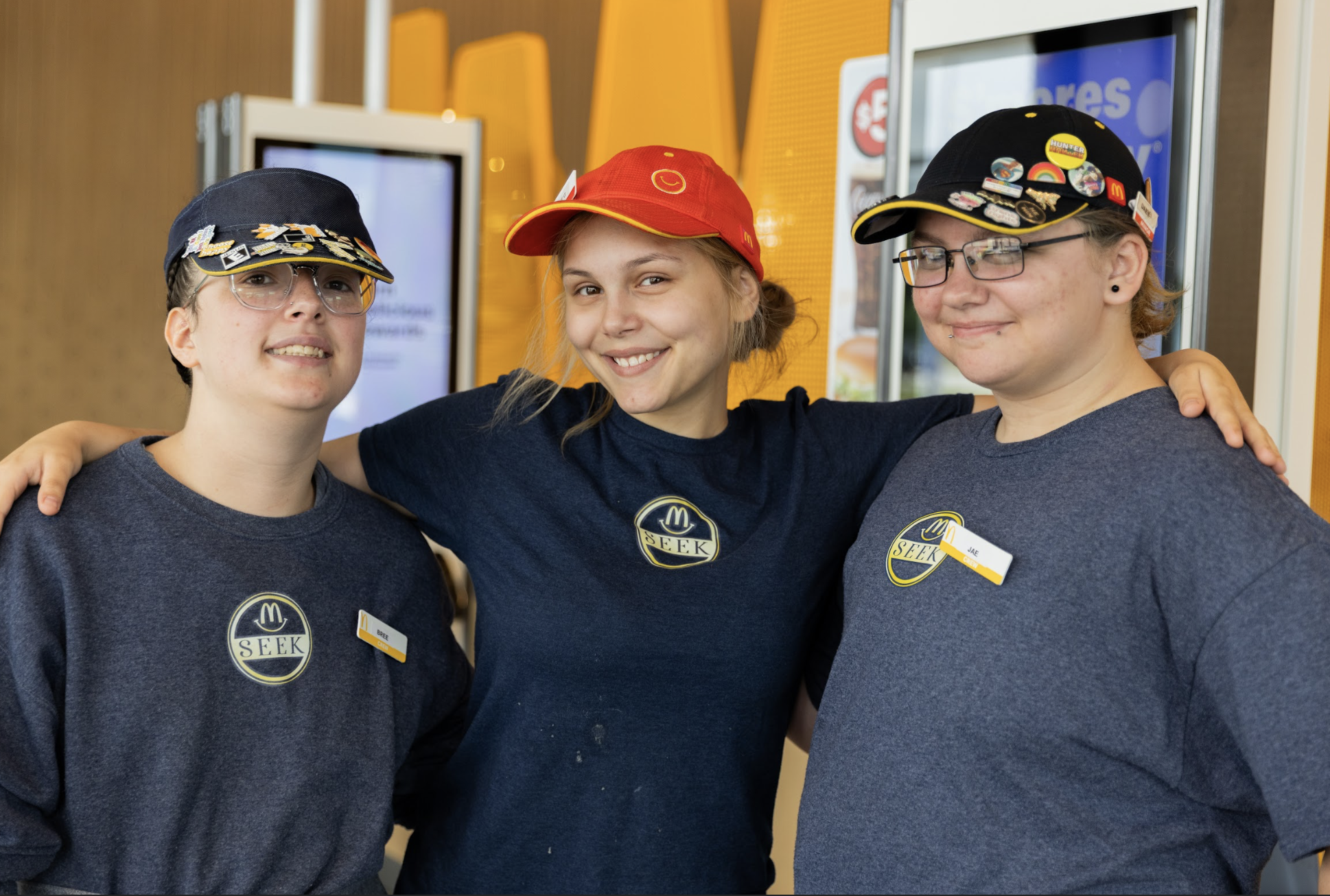 Three McDonald's employees, two women and one girl, are smiling and standing together with their arms around each other in a fast food restaurant. They are wearing McDonald's uniforms with name tags and caps with various pins.