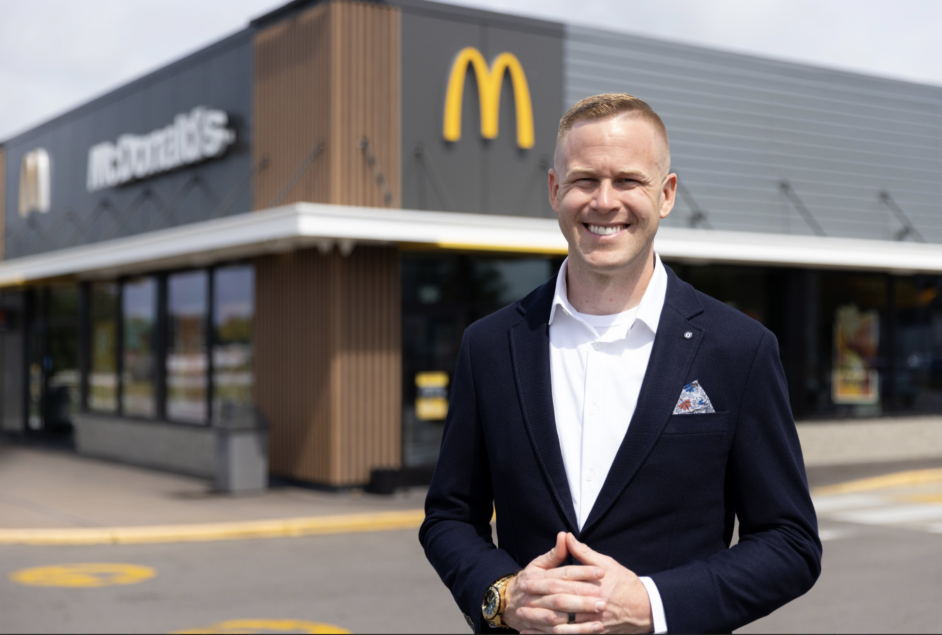 A man in a navy blazer and white shirt smiling with hands clasped in front, standing outside a McDonald's restaurant with modern design and golden arches logo in the background.