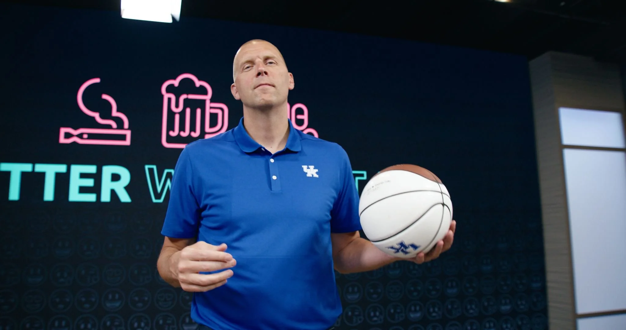 A man in a blue polo shirt with a Kentucky Wildcats logo holding a basketball in a room with a dark wall and neon signs, including a beer mug, a cigarette, and the word 'TWEETER'.